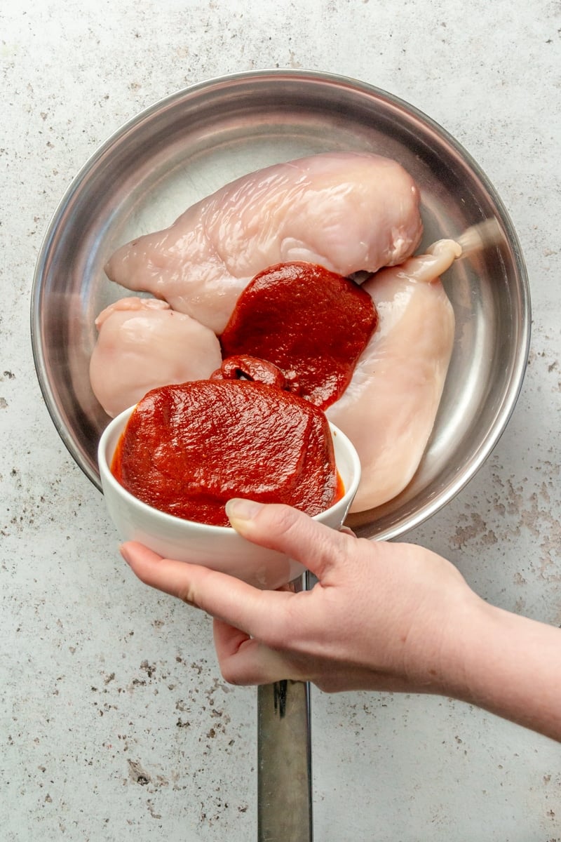 A red colored sauce is shown beeping poured over the top of three chicken breasts which sit in a metal skillet.