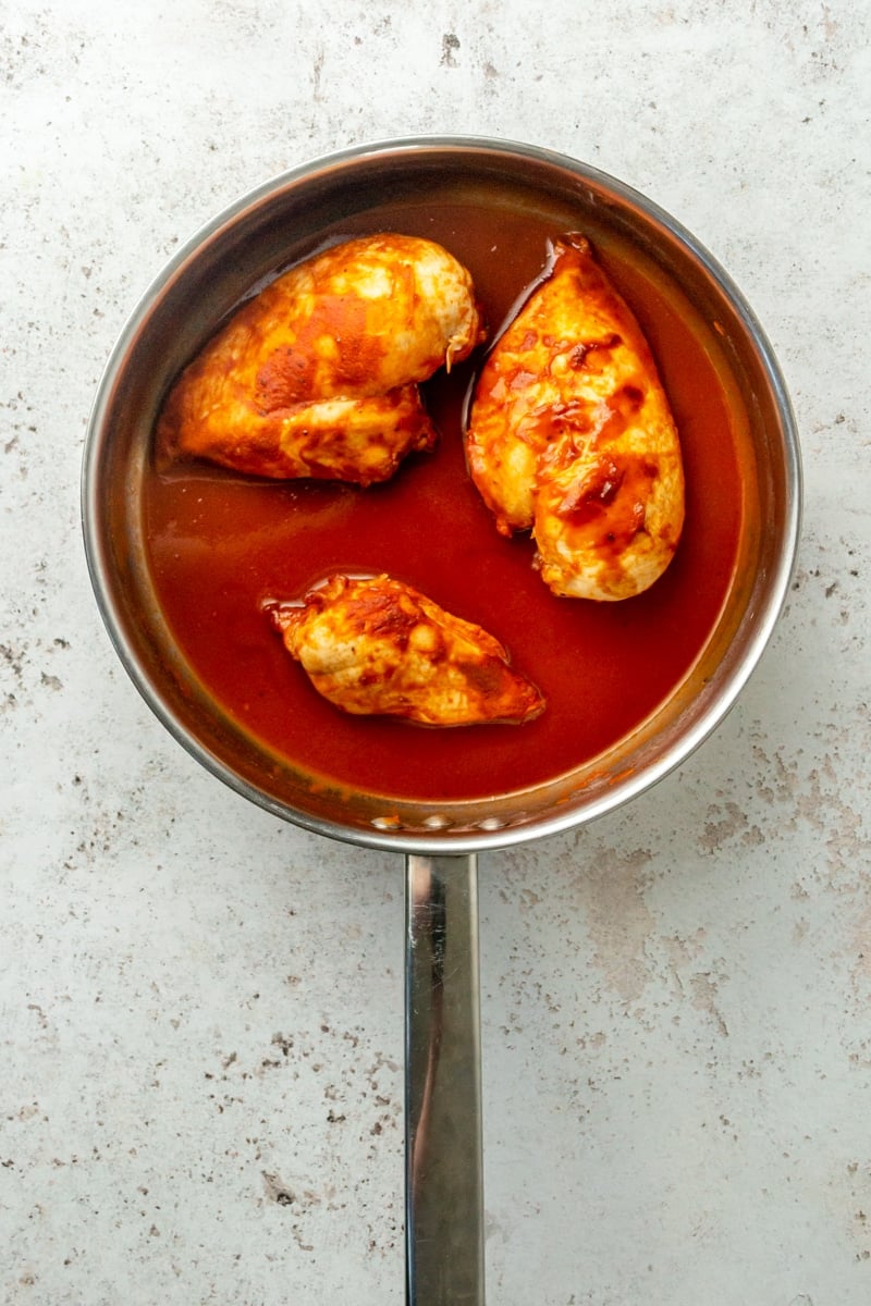 Chicken breasts sit in a red sauce in a metal skillet.