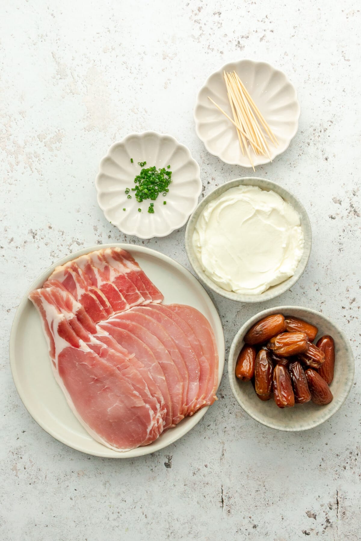 Ingredients for bacon wrapped stuffed dates sit in a variety of bowls and plates on a light grey surface.