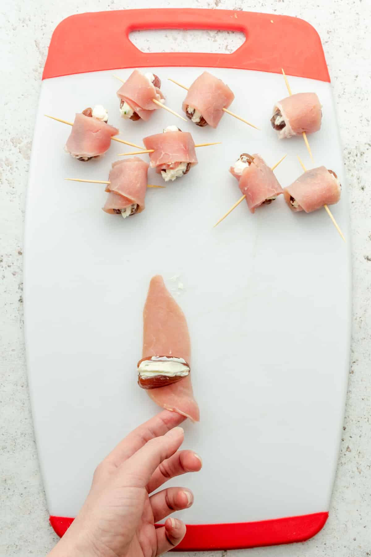 A cream cheese stuffed date has a bacon strip rolled around it on a white plastic chopping board on a light gray surface.