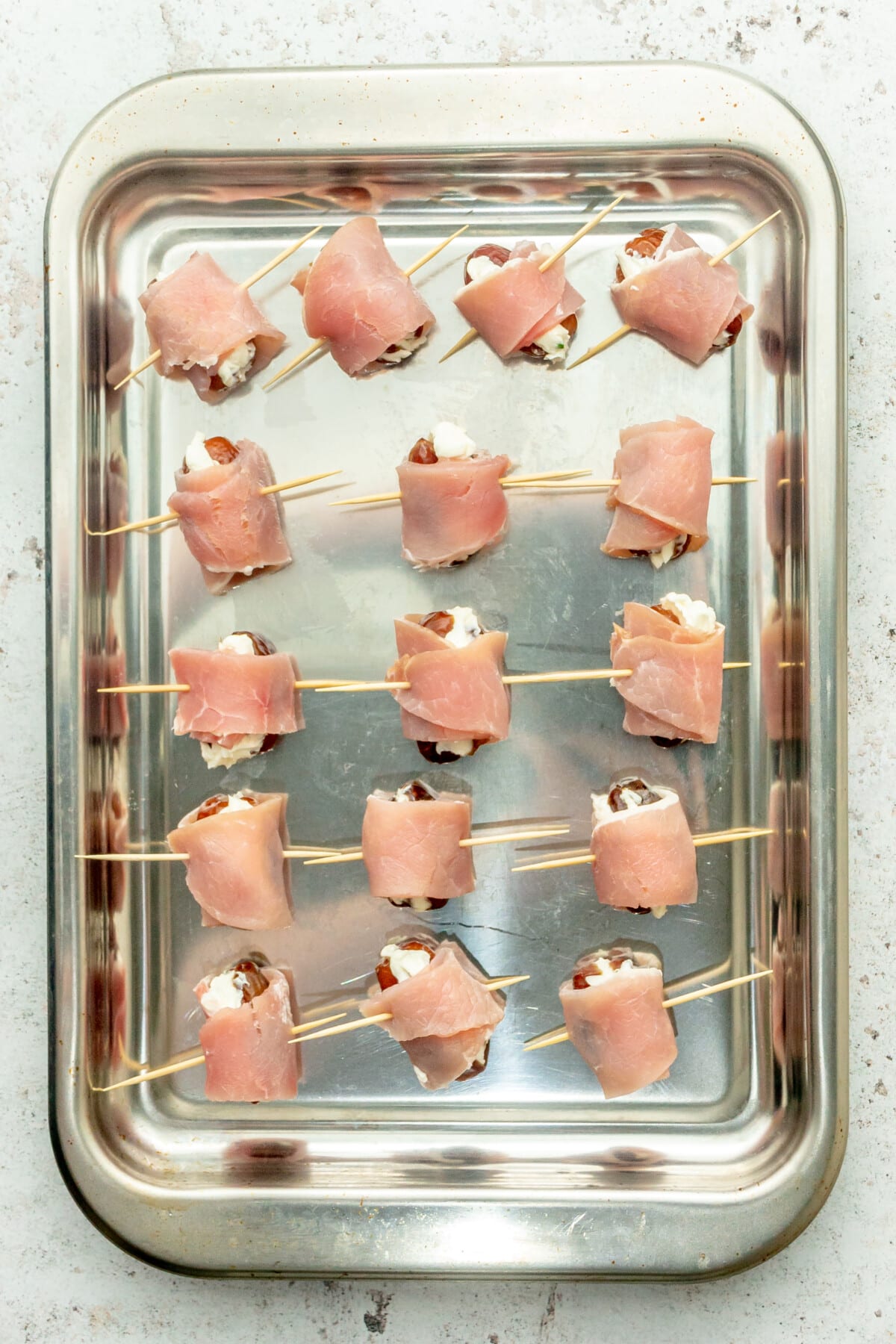 Bacon wrapped cream cheese stuffed dates pierced with toothpicks sit on a rimmed stainless steel tray on a light gray surface.