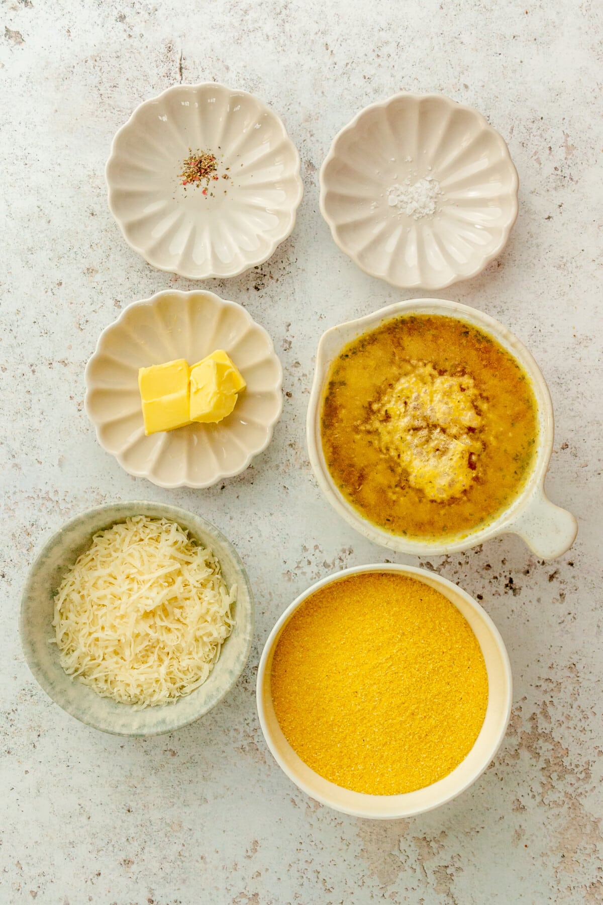 Ingredients for basic no stir polenta sit in a variety of bowls and plates on a light grey surface.