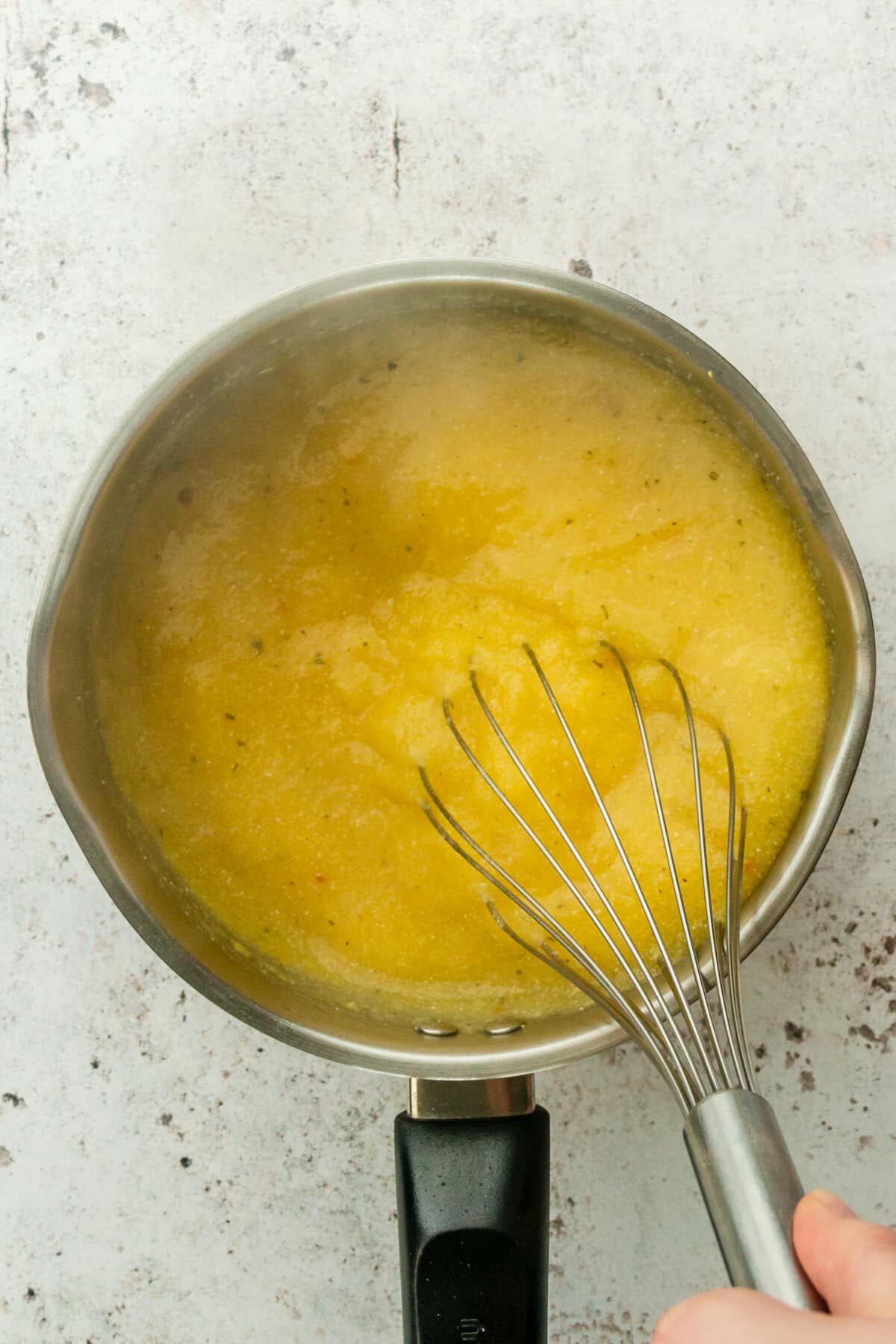 Polenta is whisked in a stainless steel saucepan on a light grey surface.