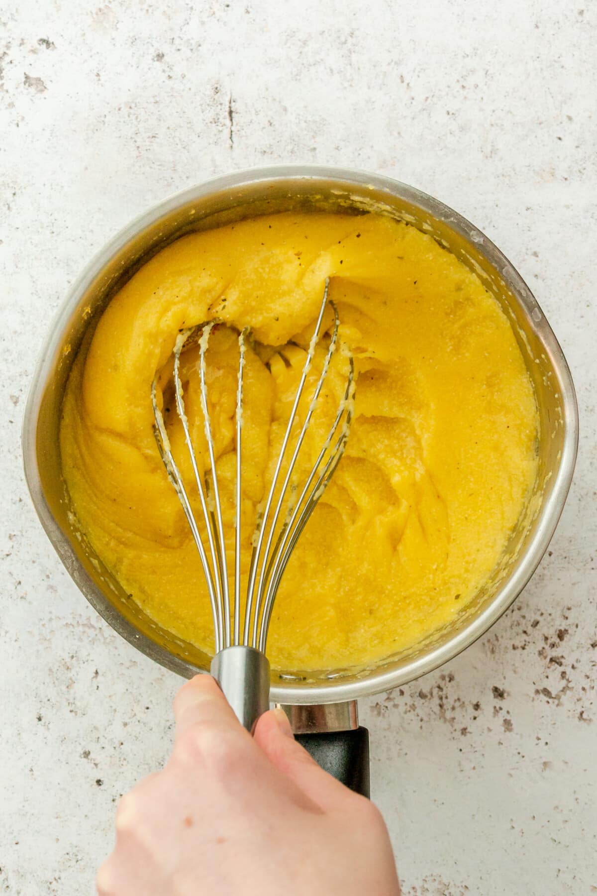 Cooked thick polenta is whisked in a stainless steel saucepan on a light grey surface.