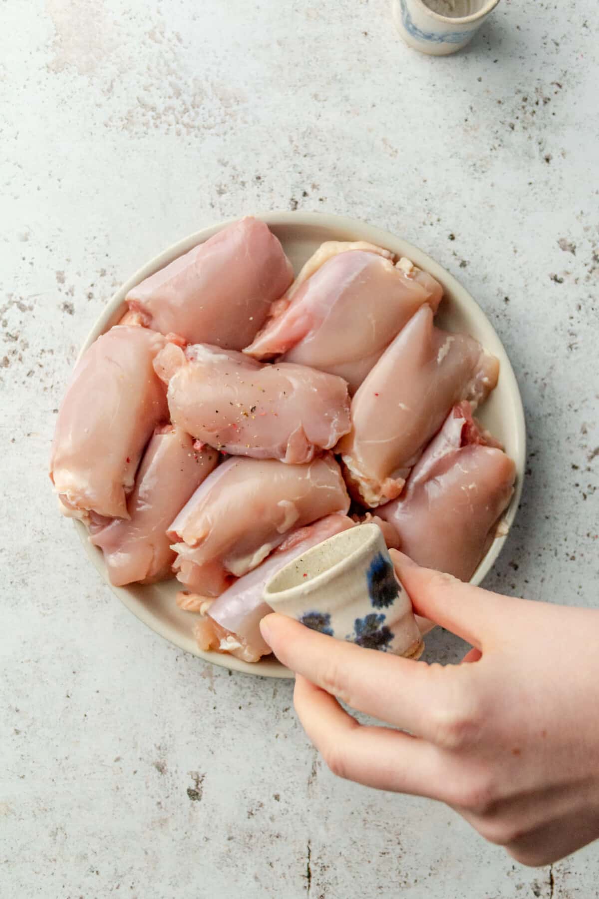 A person sprinkling salt and pepper onto a plate of raw chicken thighs.