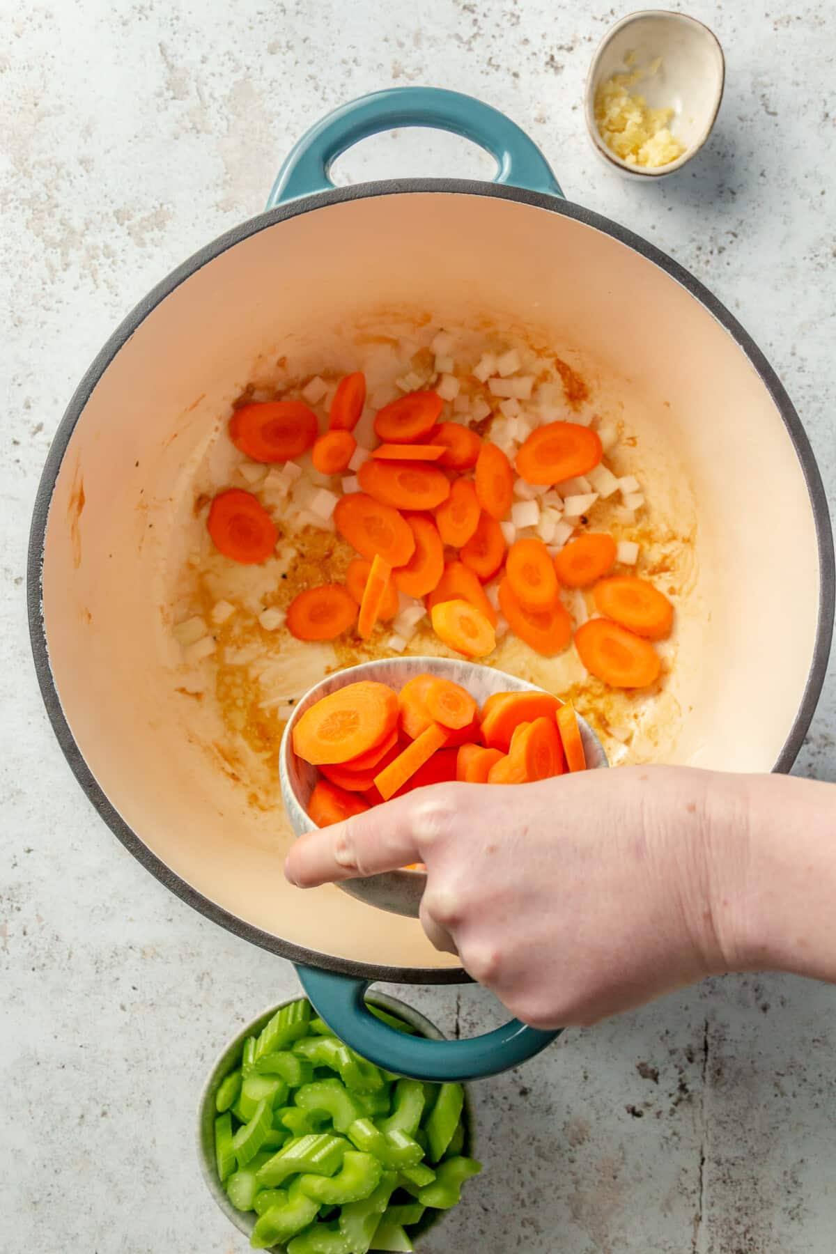 A person adding a small bowl of coined carrots into an enameled cast iron pot to make chicken noodle soup.