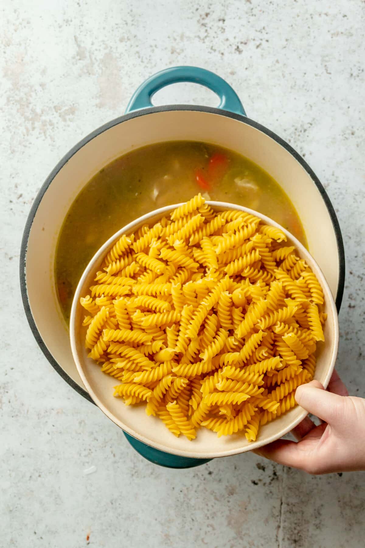 A person holding a bowl of uncooked rotini over a pot of chicken noodle soup.