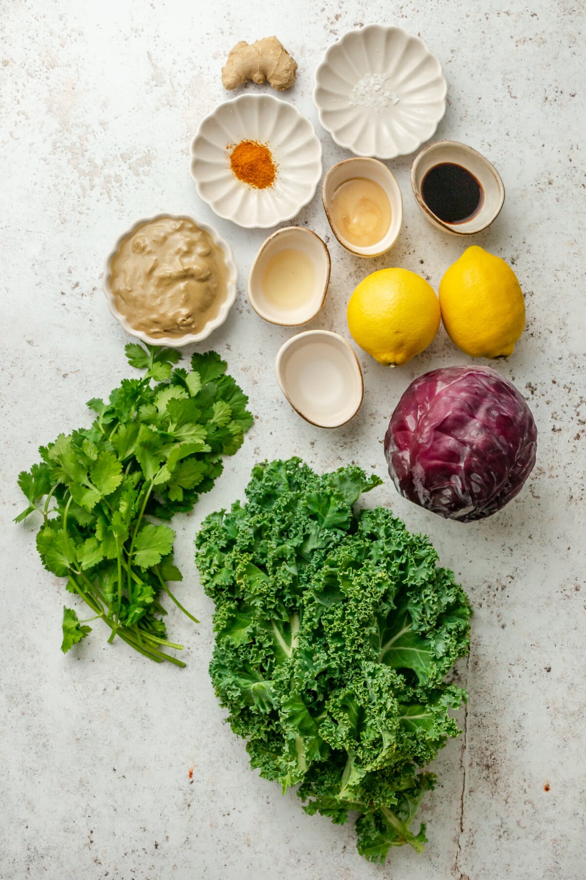Ingredients for a big batch kale salad sit in a variety of bowls on a light grey surface.