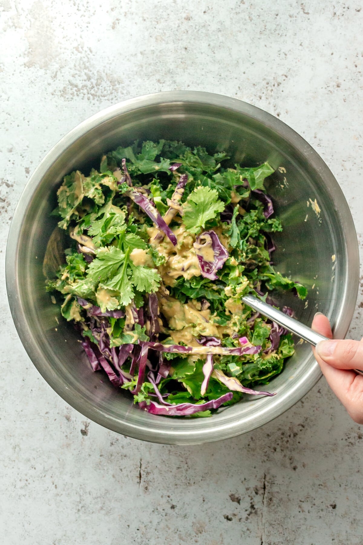 A dressing is stirred through a kale salad in a stainless steel bowl on a light grey surface.