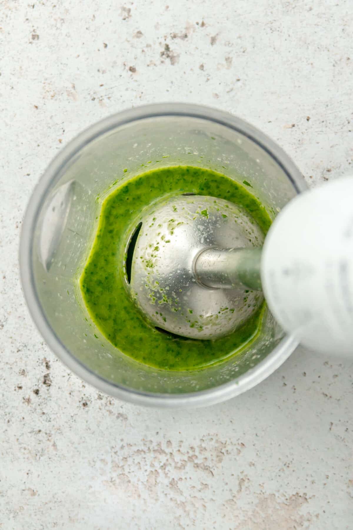 A coriander lime sauce is shown in a blender beaker with an immersion blender on a light grey colored surface.
