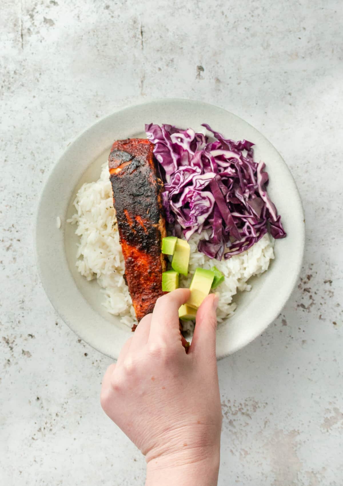 A cube of avocado is shown being placed on top of coconut rice beside blackened salmon and shredded purple cabbage on a light grey colored surface.