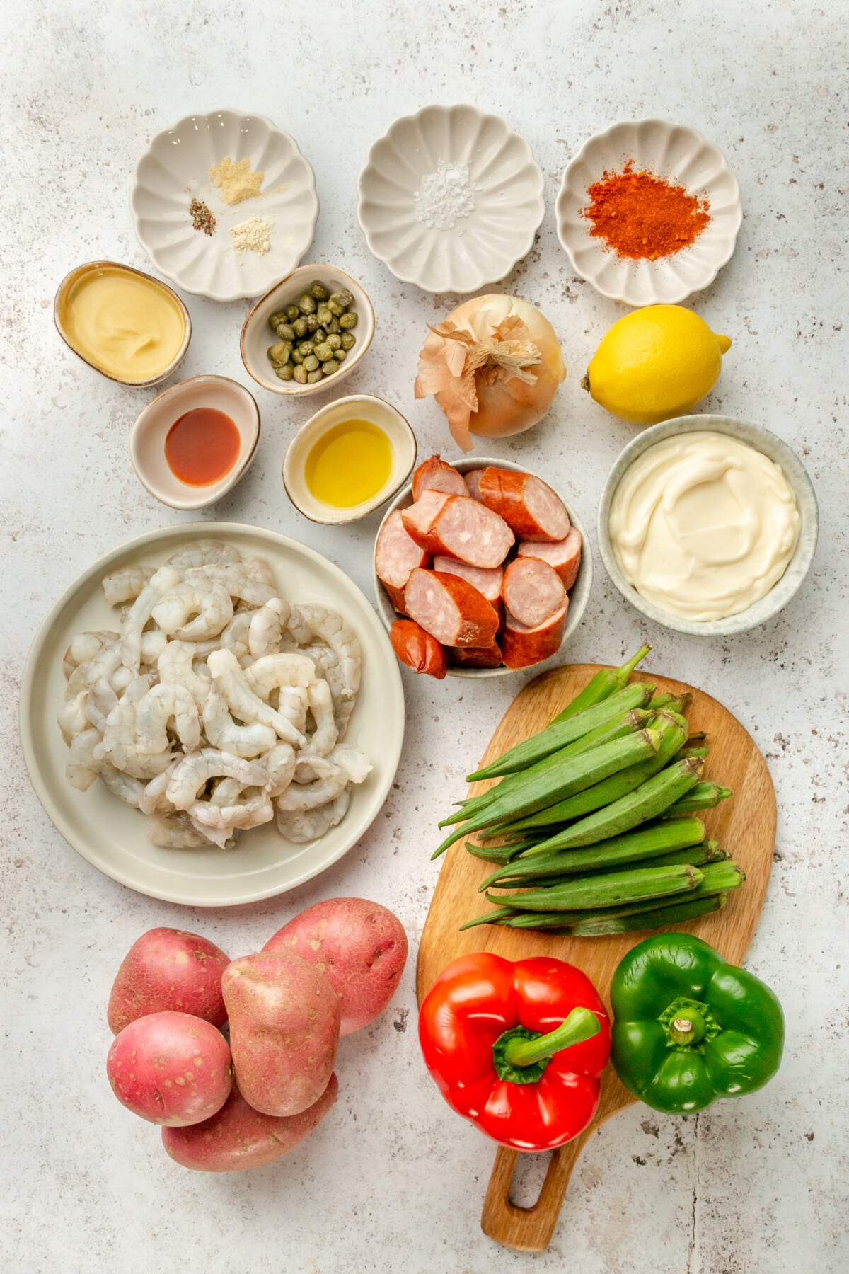 Ingredients for cajun shrimp sheet pan dinner sit in a variety of bowls and plates on a light grey surface.