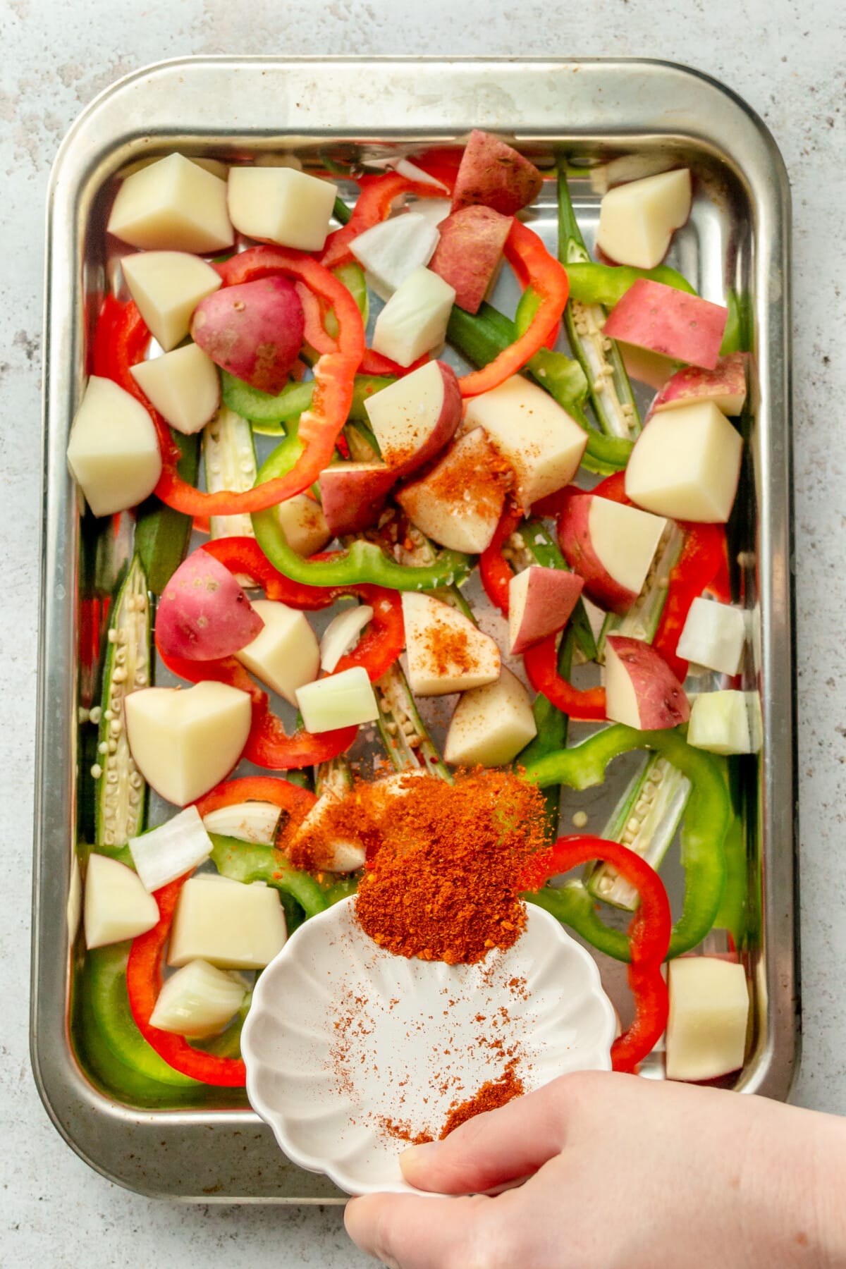 Cajun seasoning is tossed over cut vegetables sitting on a stainless steel rimmed baking sheet on a light grey surface.