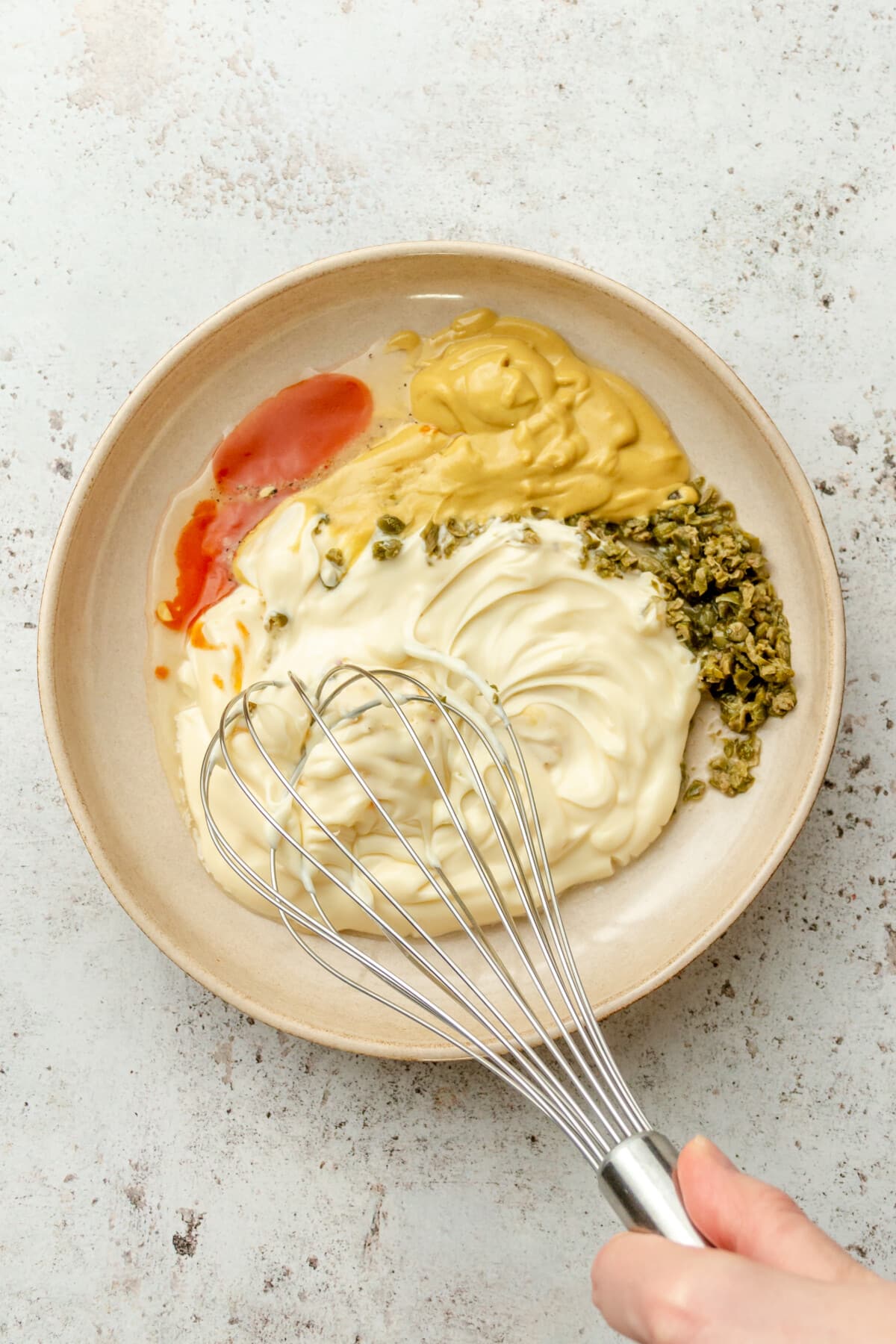 A remoulade sauce is whisked in a ceramic shallow bowl on a light grey surface.