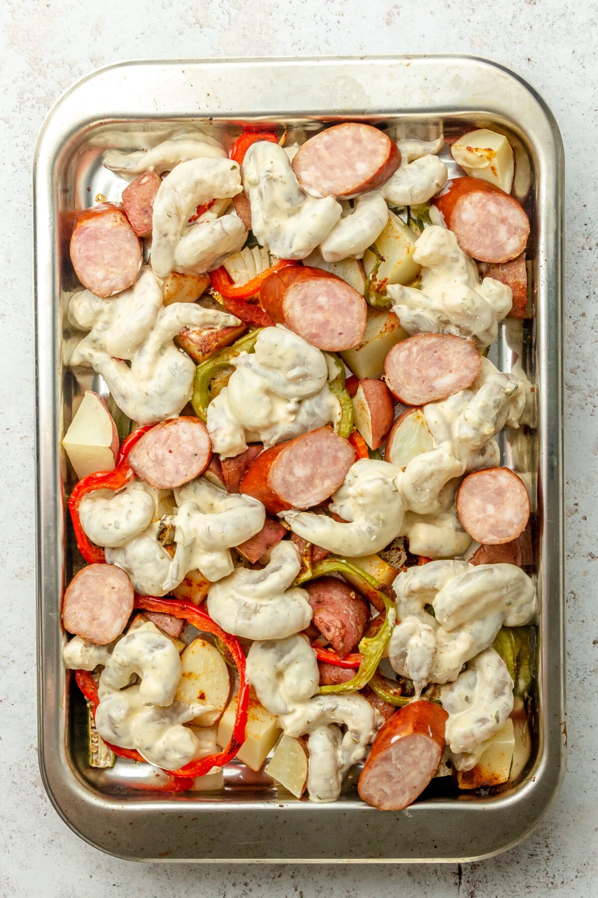 Shrimp coated in a remoulade sauce and andouille sausage pieces sit on top of roasted vegetables on a rimmed stainless steel baking sheet on a light grey surface.