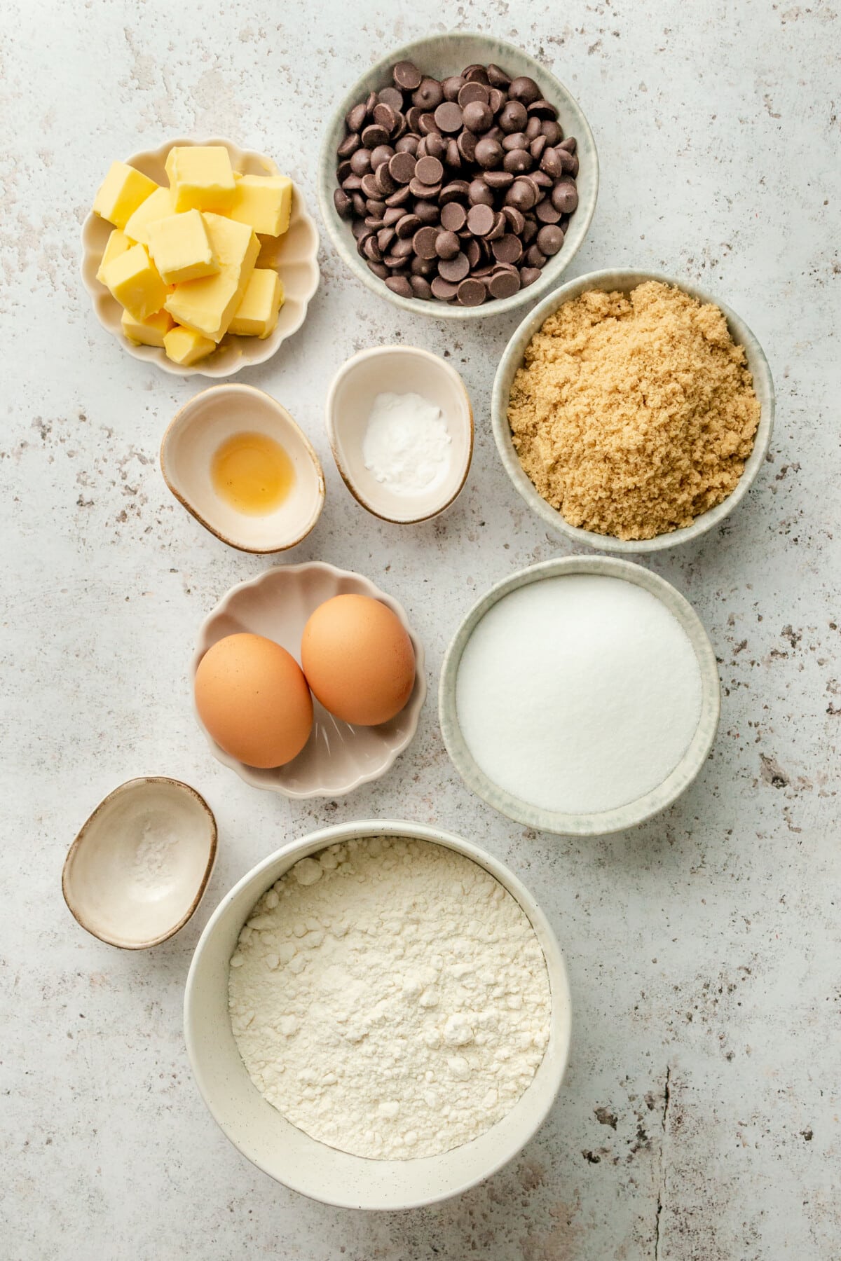 Ingredients for gluten free chocolate chip cookies sit in a variety of bowls and plates on a light grey surface.