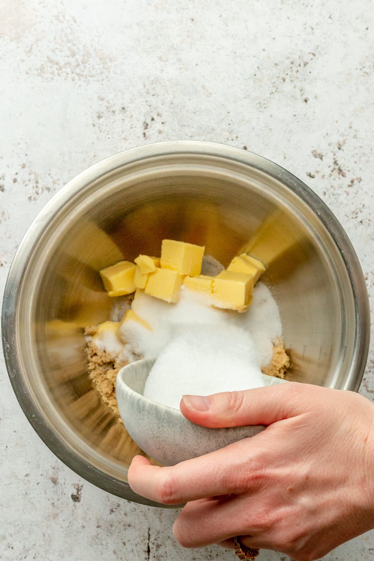 Sugar is tossed over butter in a stainless steel bowl on a light grey surface.