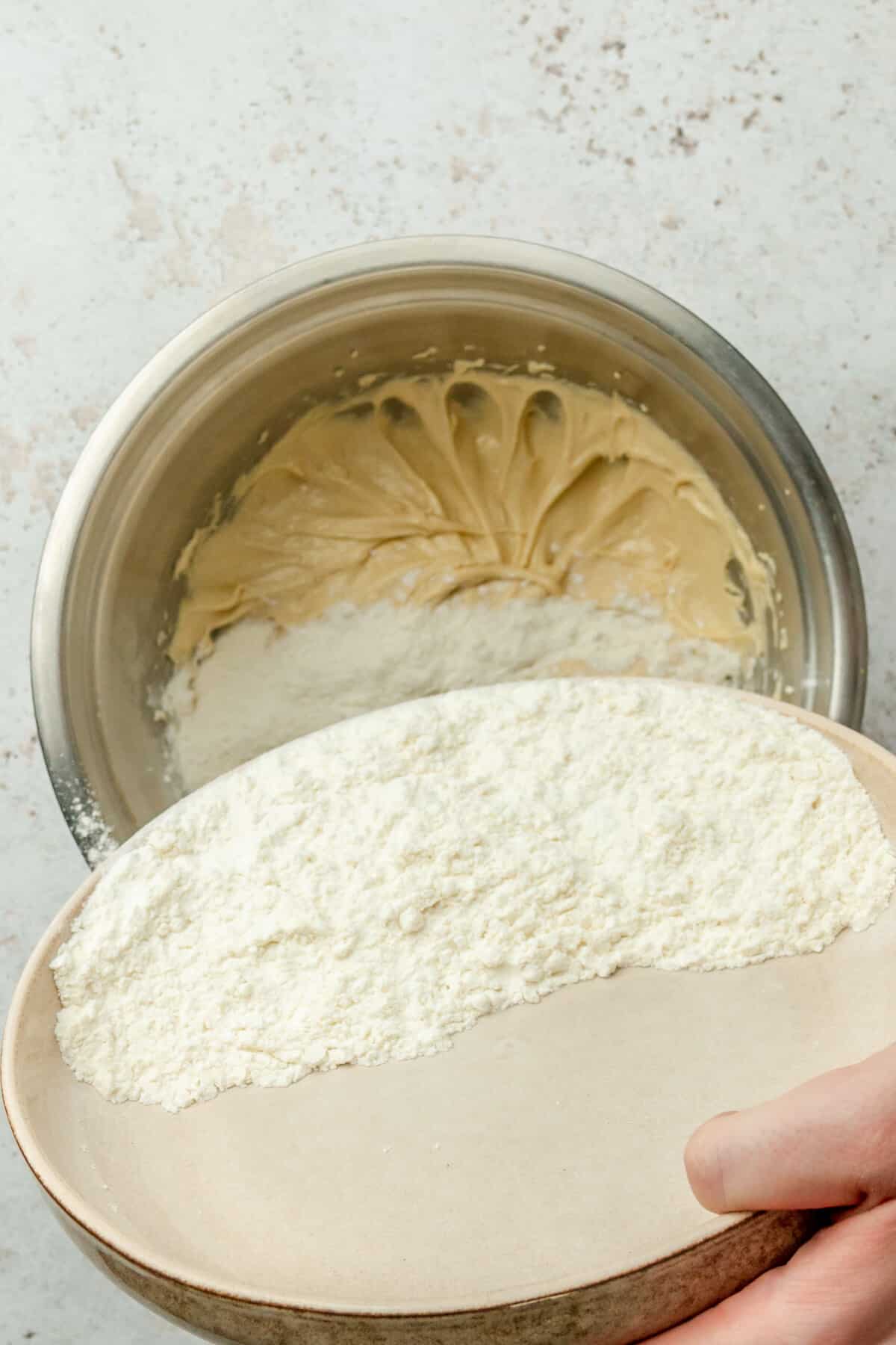 A portion of dry ingredients are tossed over a butter and egg cookie mixture in a stainless steel bowl on a light grey surface.