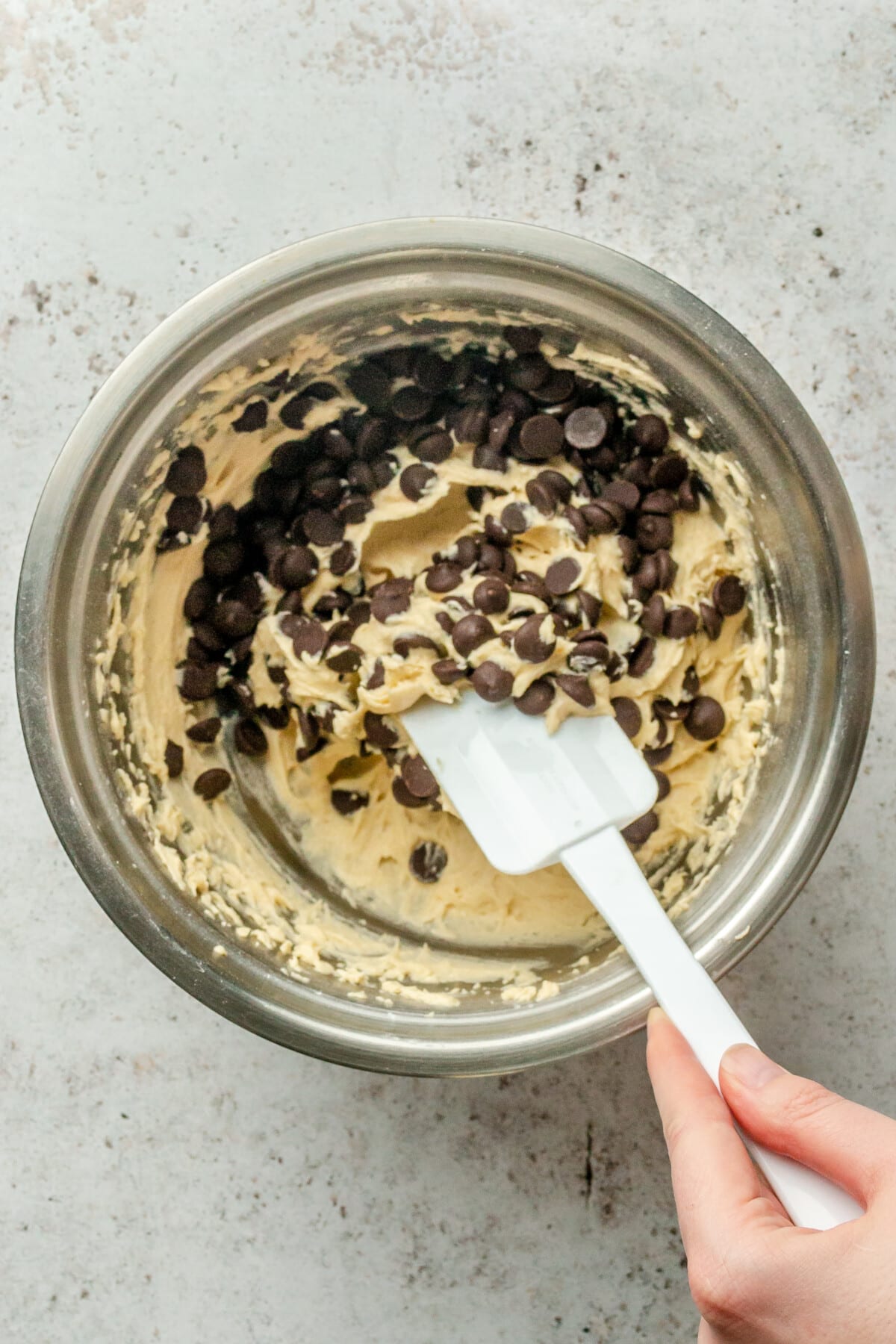 Dark chocolate chips are stirred through cookie batter in a stainless steel bowl on a light grey surface.