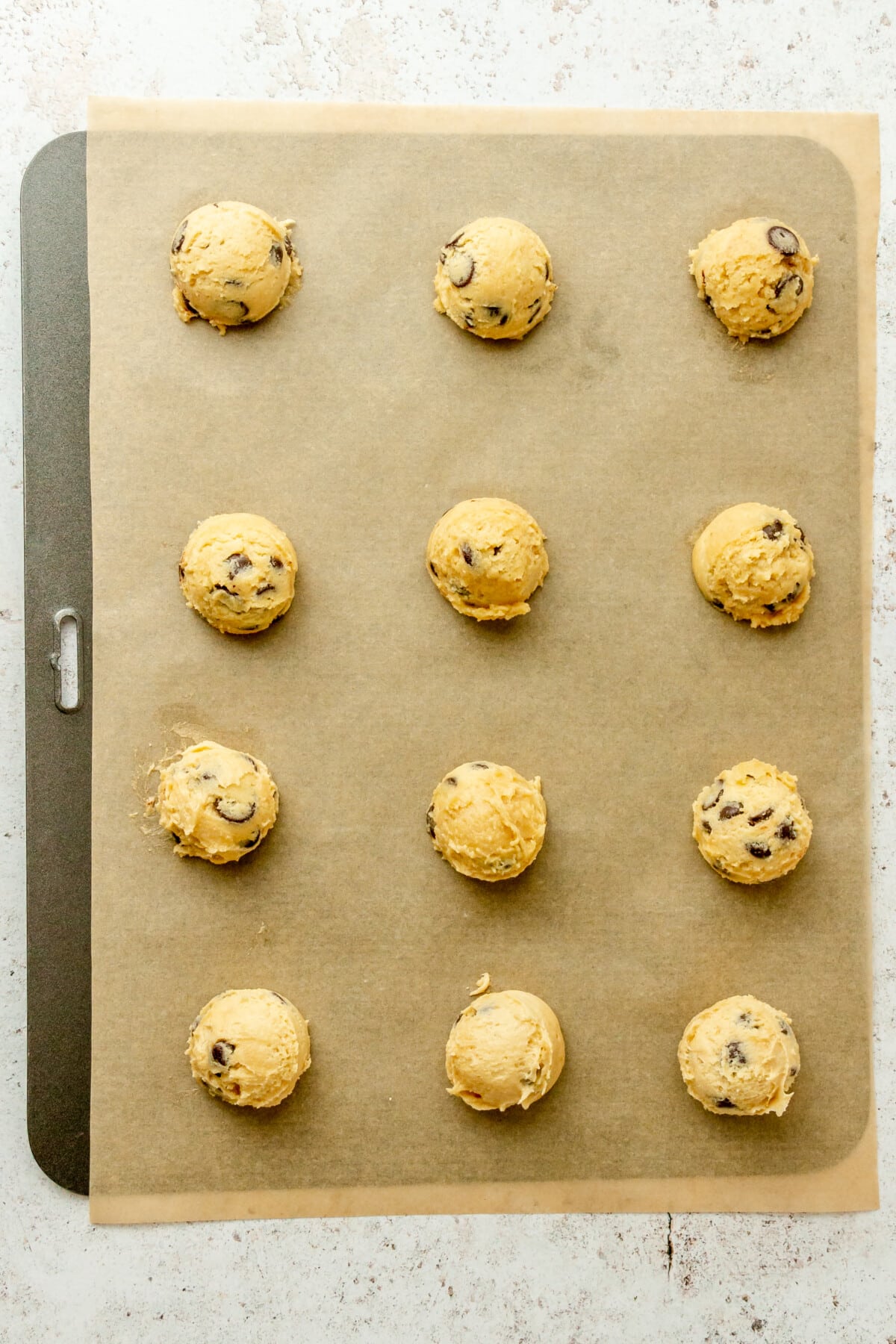 Scoops of gluten free chocolate chip cookie dough sit on a lined baking tray on a light grey surface.