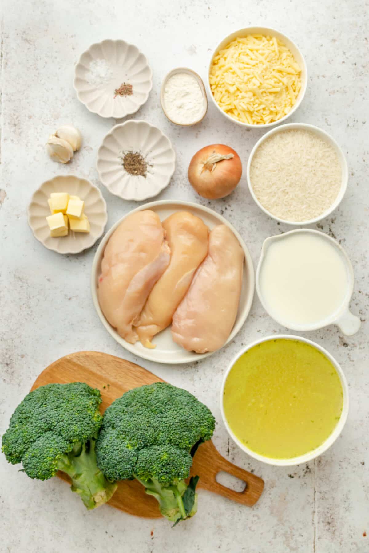 Ingredients for chicken broccoli and rice casserole sit in a variety of bowls and plates on a light grey surface.