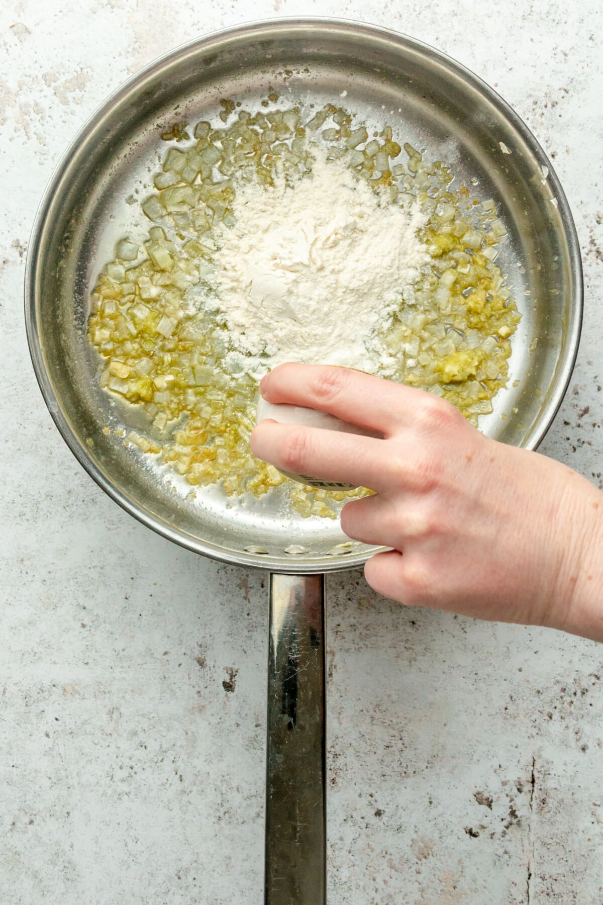 Flour is tossed over melted butter and translucent cubed onion in a stainless steel saucepan on a light grey surface.
