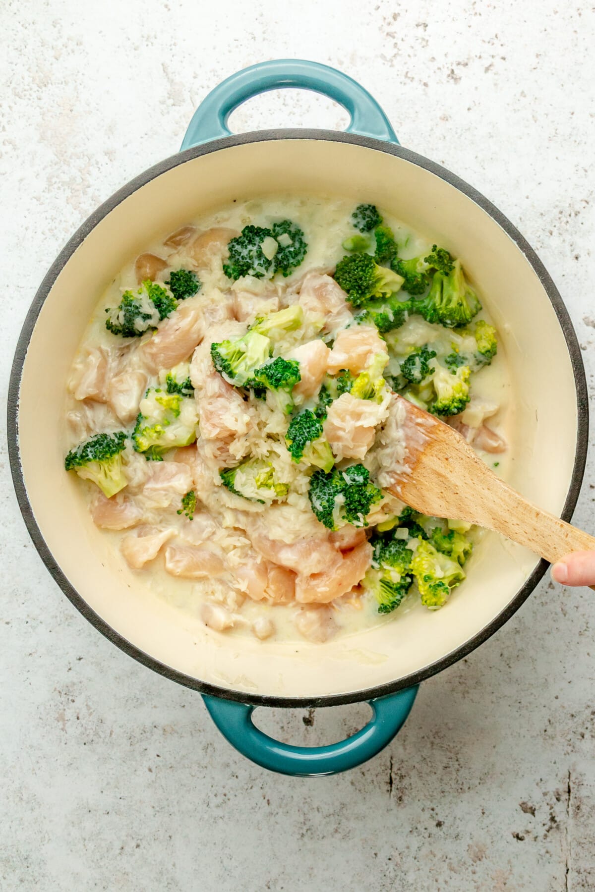 Rice, broccoli florets and chicken cubes are stirred in a Dutch oven on a light grey surface.