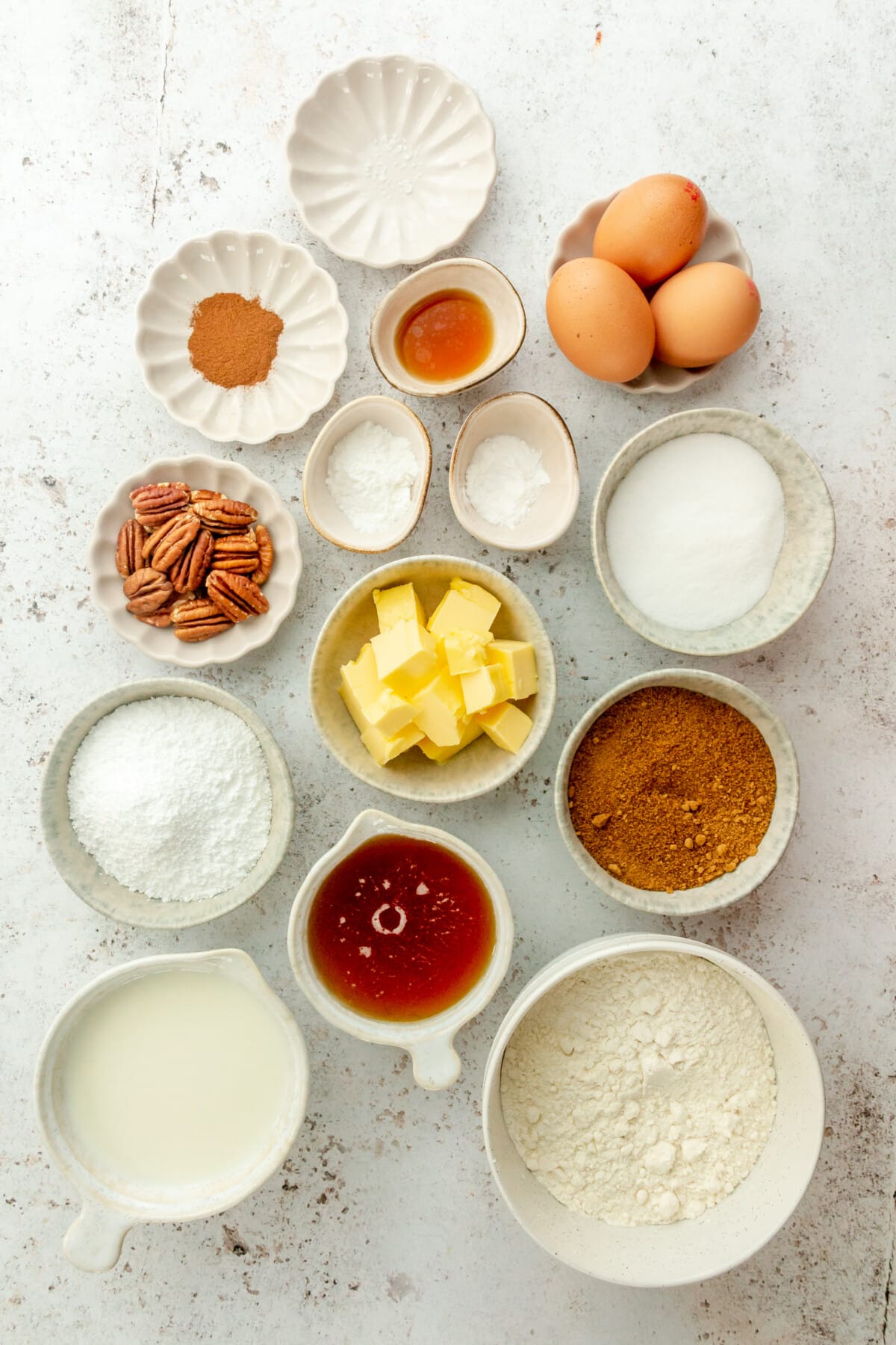 Ingredients for cinnamon roll coffee cake sit in a variety of bowls and plates on a light grey surface.