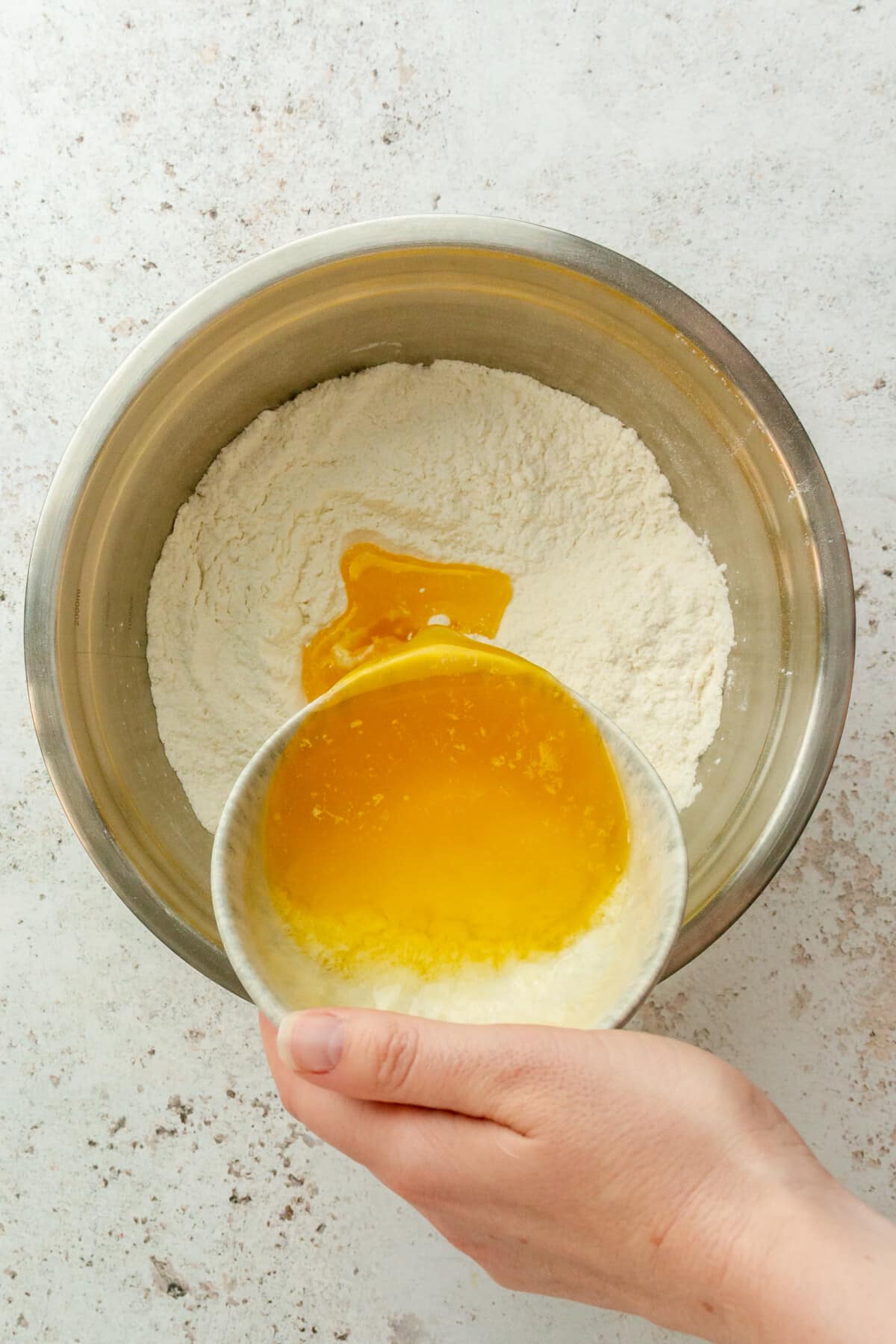 Melted butter is poured over a flour mixture in a stainless steel bowl on a light grey surface.