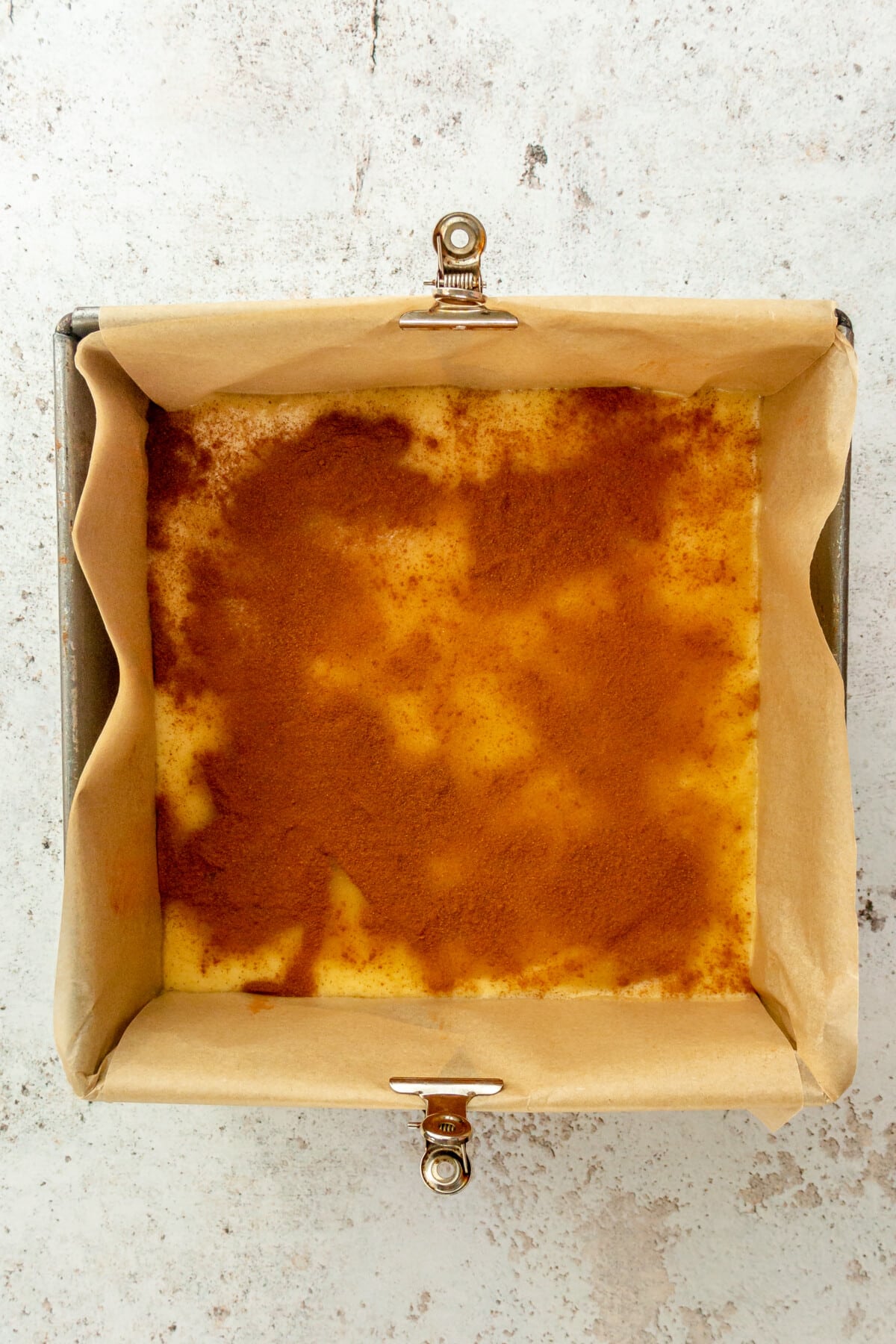 A cinnamon sugar mixture sits on top of a cinnamon roll coffee cake mixture in a square metal tin lined with parchment paper on a light grey surface.