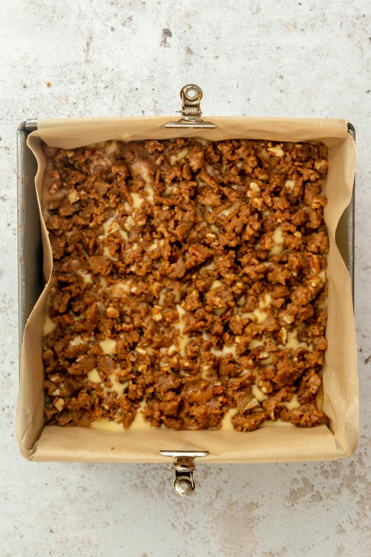 A cinnamon roll coffee crumb cake mixture sits in a square metal tin prior to baking on a light grey surface.