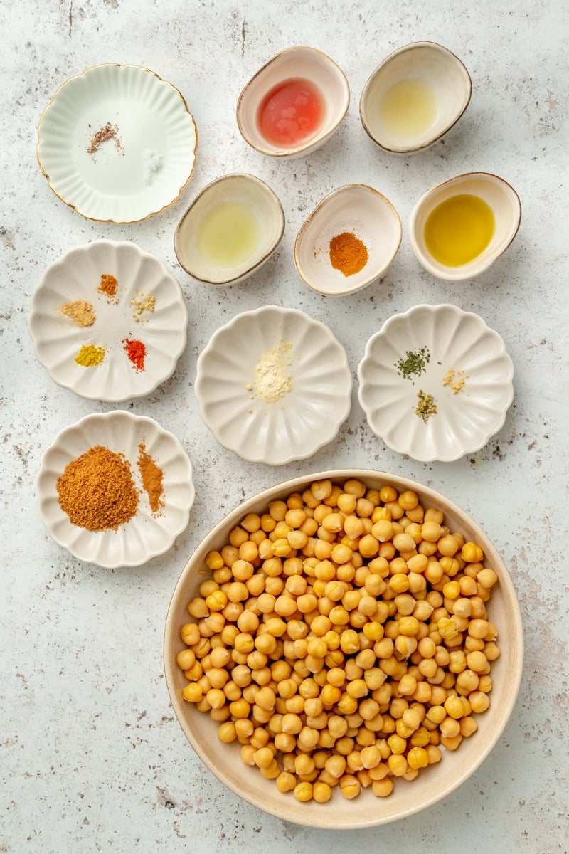 Ingredients to make crispy roasted chickpeas sit in a variety of bowls on a light grey colored surface.