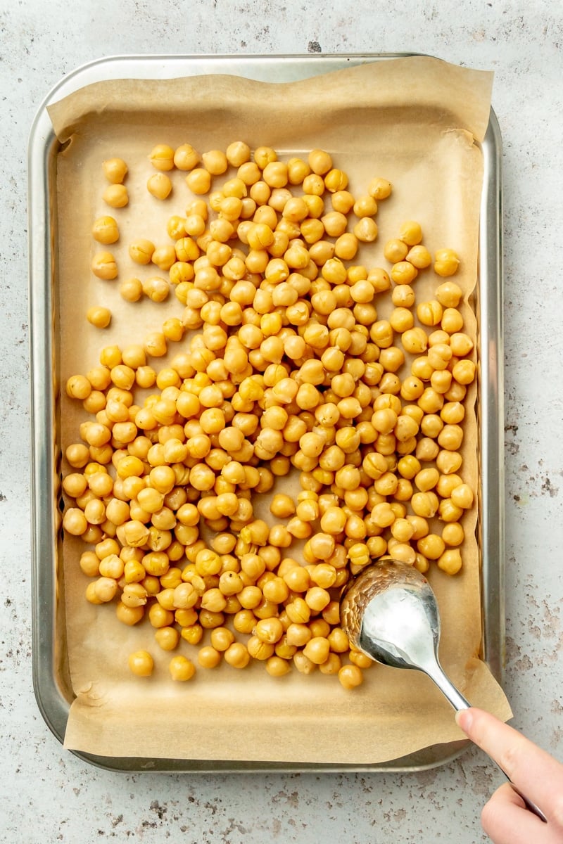 Chickpeas sit on a parchment lined baking sheet. A metal spoon is shown moving them around so they sit in an even layer.