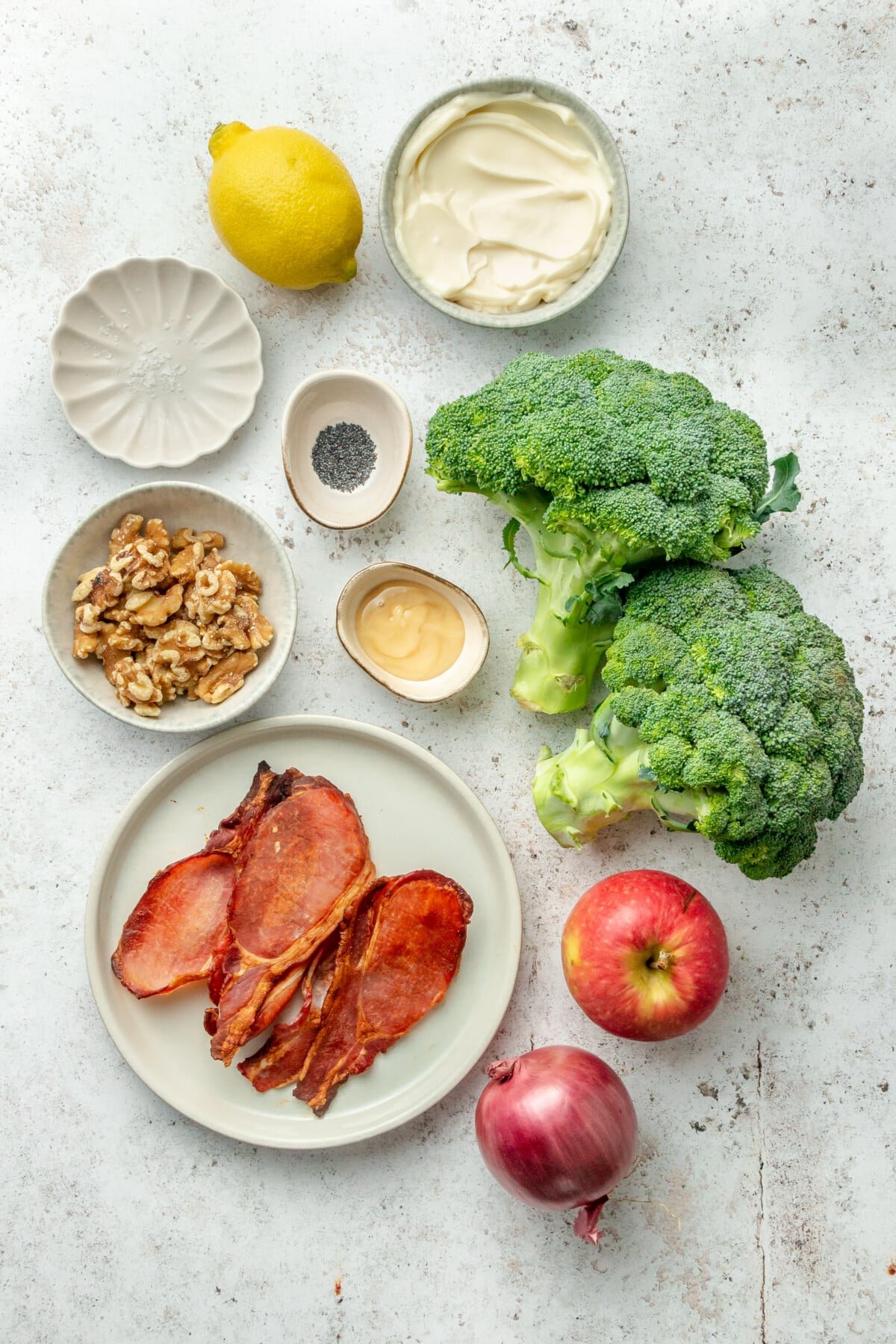 Ingredients for crunchy broccoli salad sit in a variety of plates and bowls on a light grey surface.