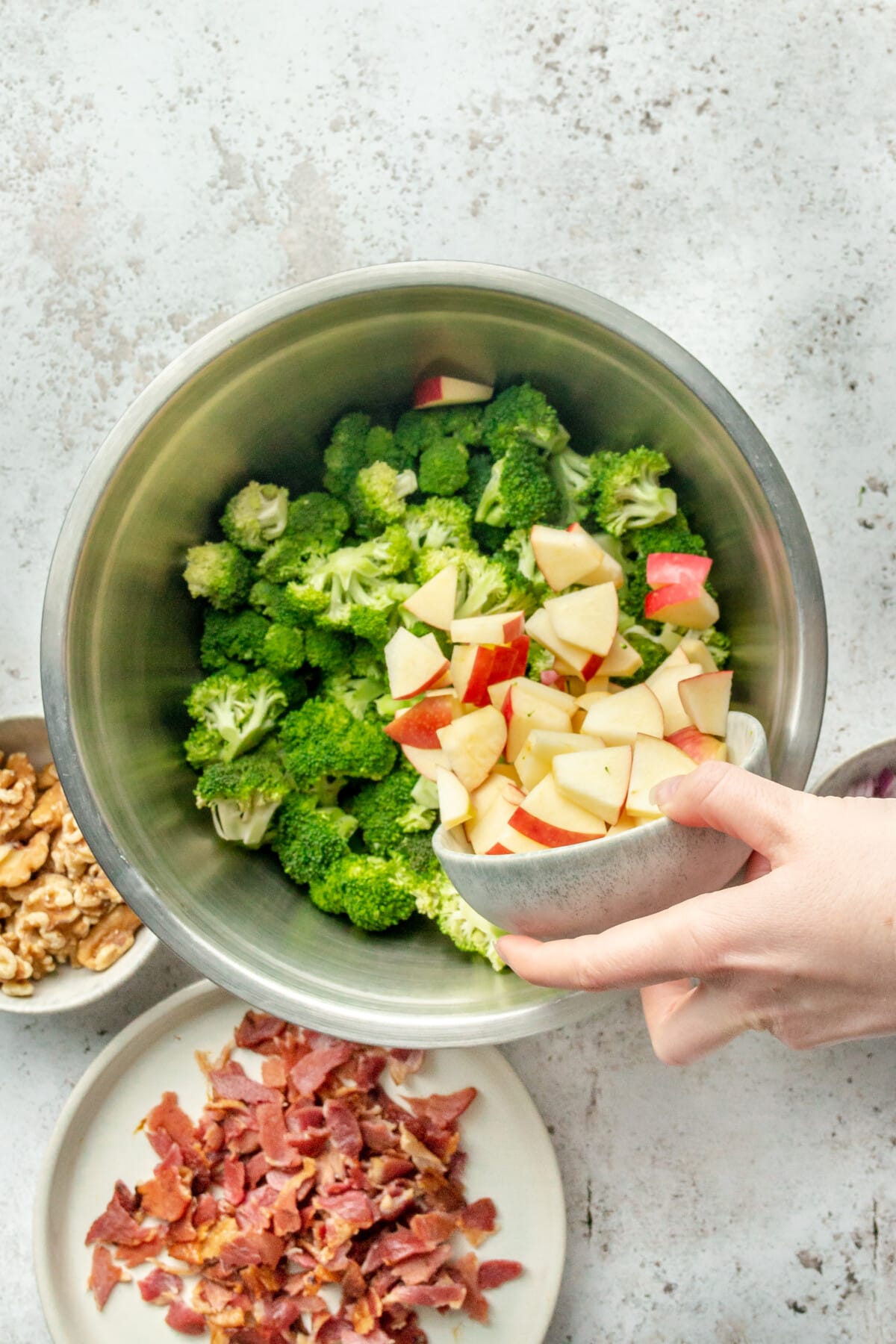 Apple chunks are tossed into a bowl with broccoli pieces for a salad in a stainless steel bowl on a light grey surface.