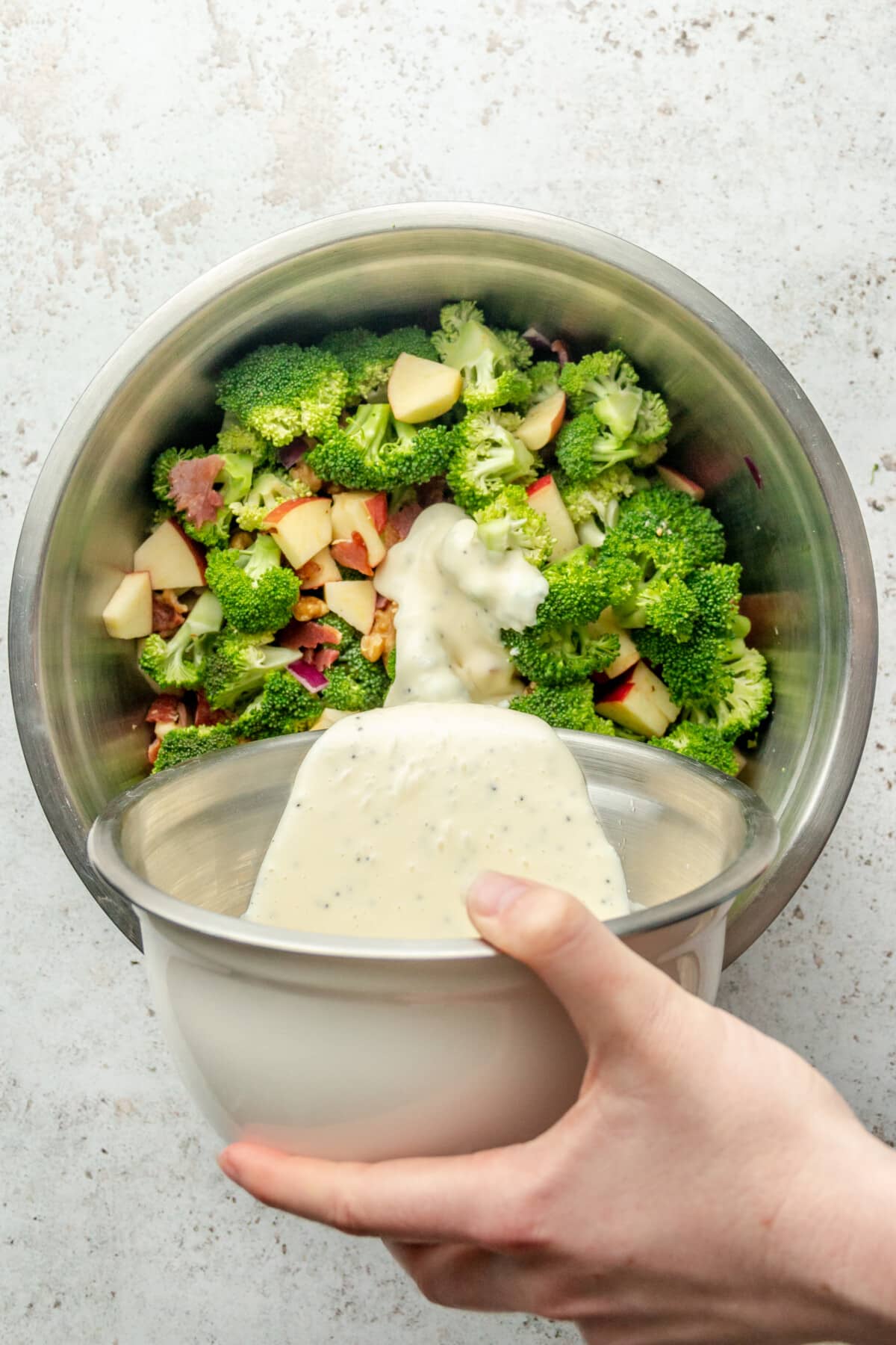 A dressing is poured over crunchy broccoli salad sitting in a stainless steel bowl on a light grey surface.
