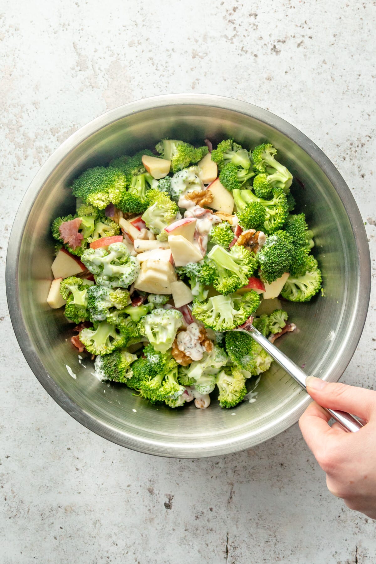 A dressing is stirred through a crunchy broccoli salad sitting in a stainless steel bowl on a light grey surface.