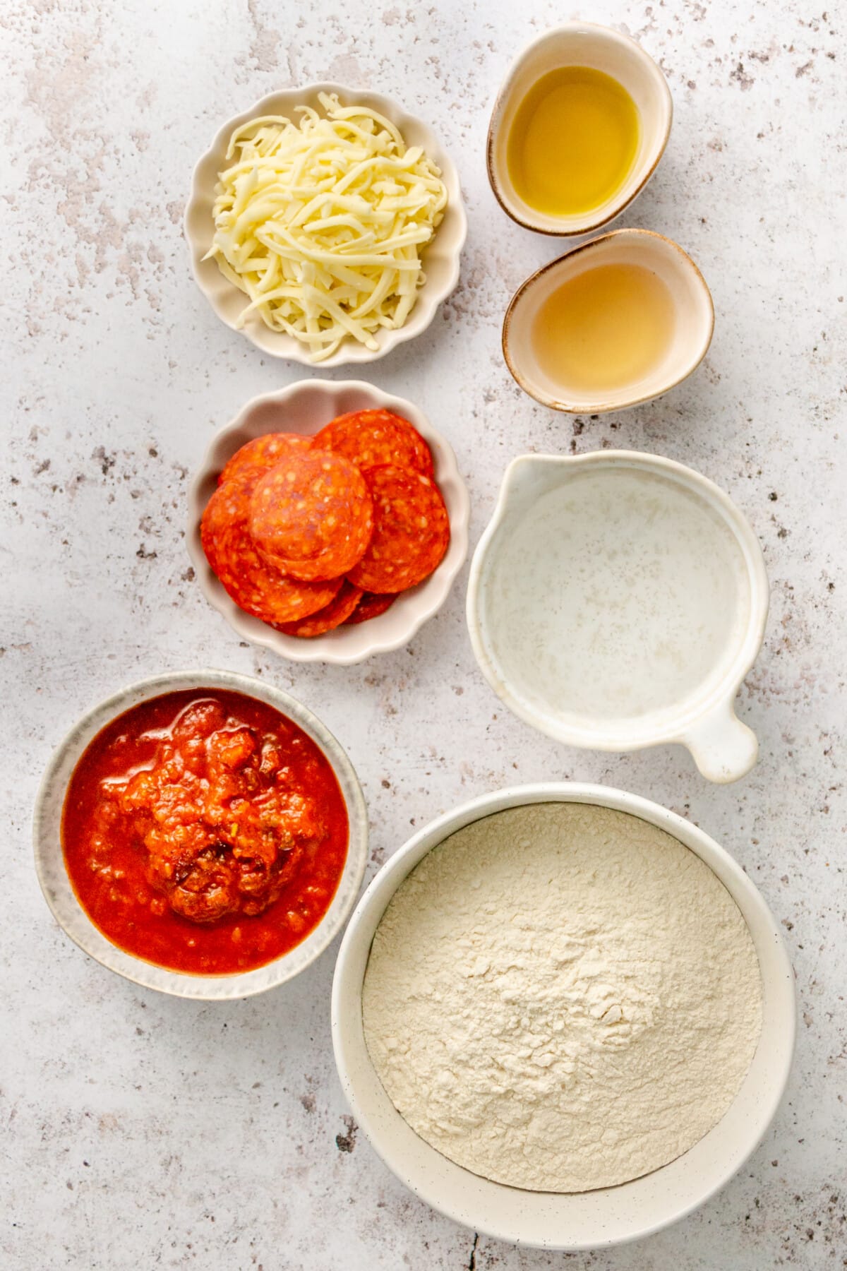 Ingredients for DIY lunchable pizza sit in a variety of plates and bowls on a light grey surface.