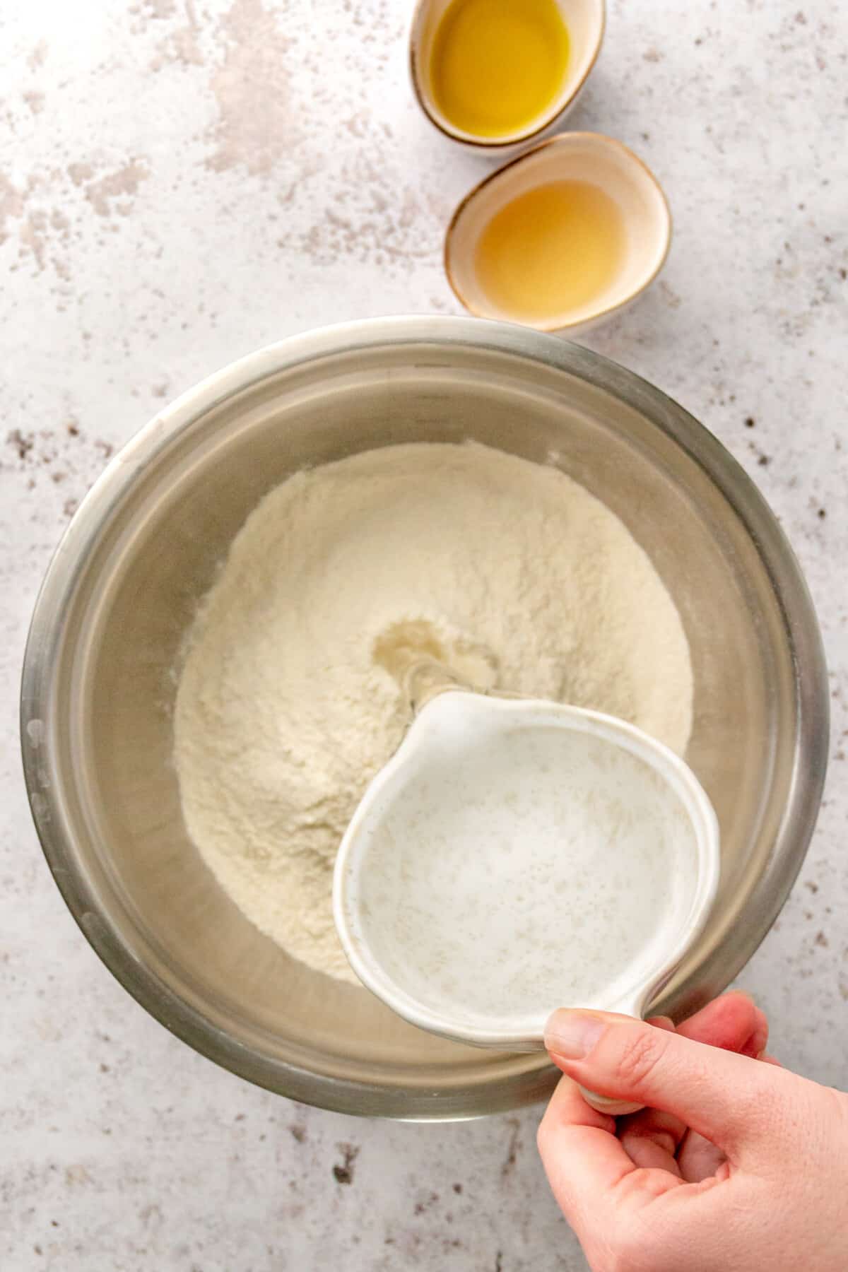 Water is poured into a pizza dough mix in a stainless steel bowl on a light grey surface.