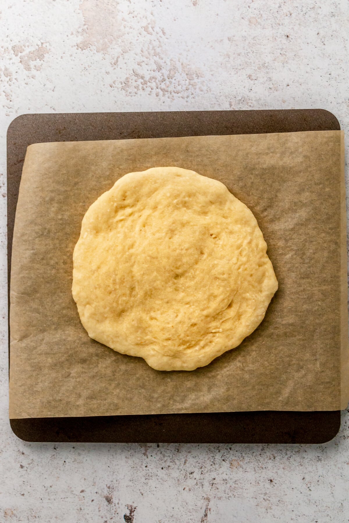 One small pizza dough sits rolled out on baking paper on a small tray on a light grey surface.