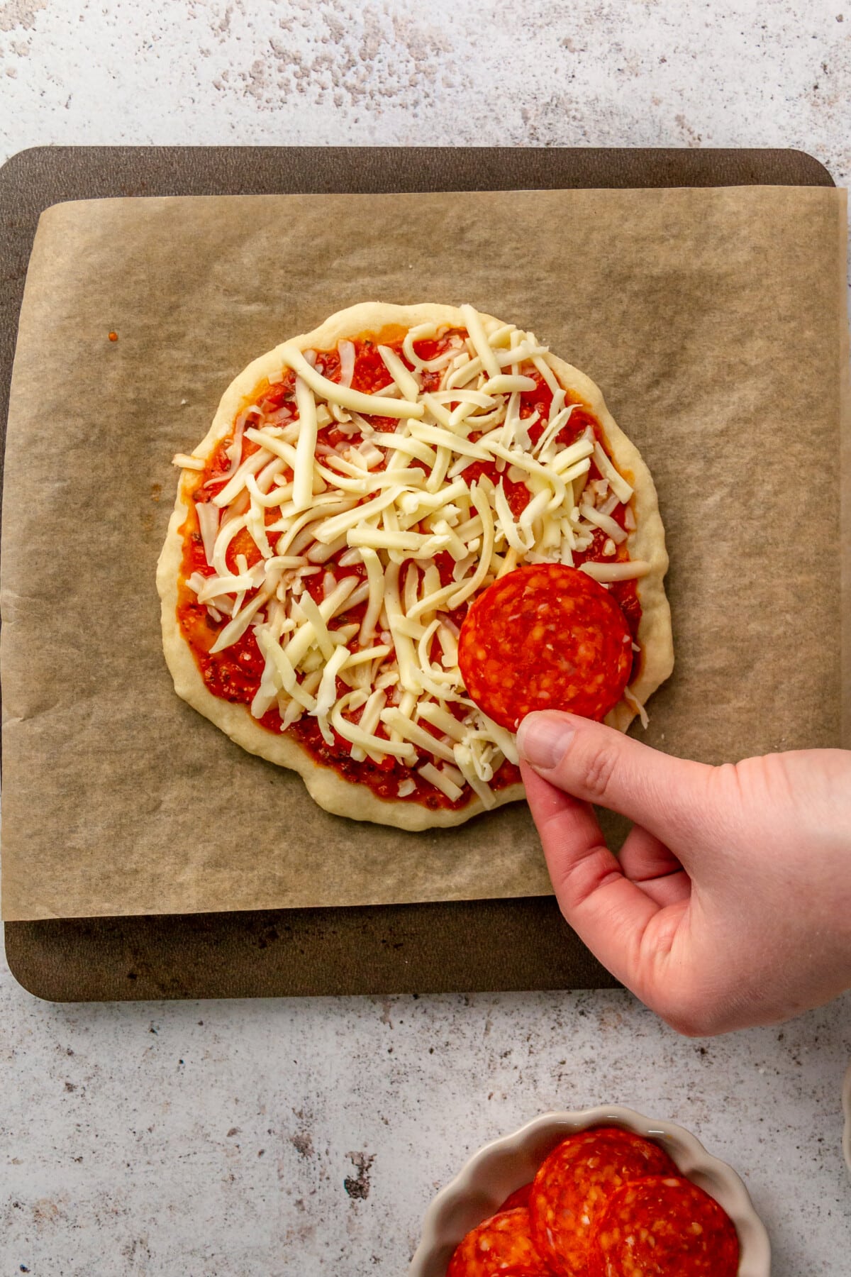 A piece of pepperoni is placed on top of shredded cheese on a small pizza sitting on baking paper on a small tray on a light grey surface.