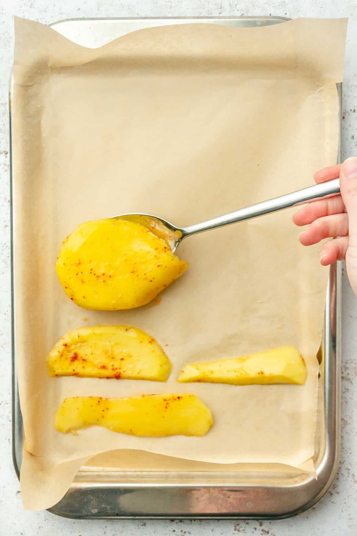 Slices of mango with spots of bright chili are laid onto a lined baking tray on a light grey colored surface.