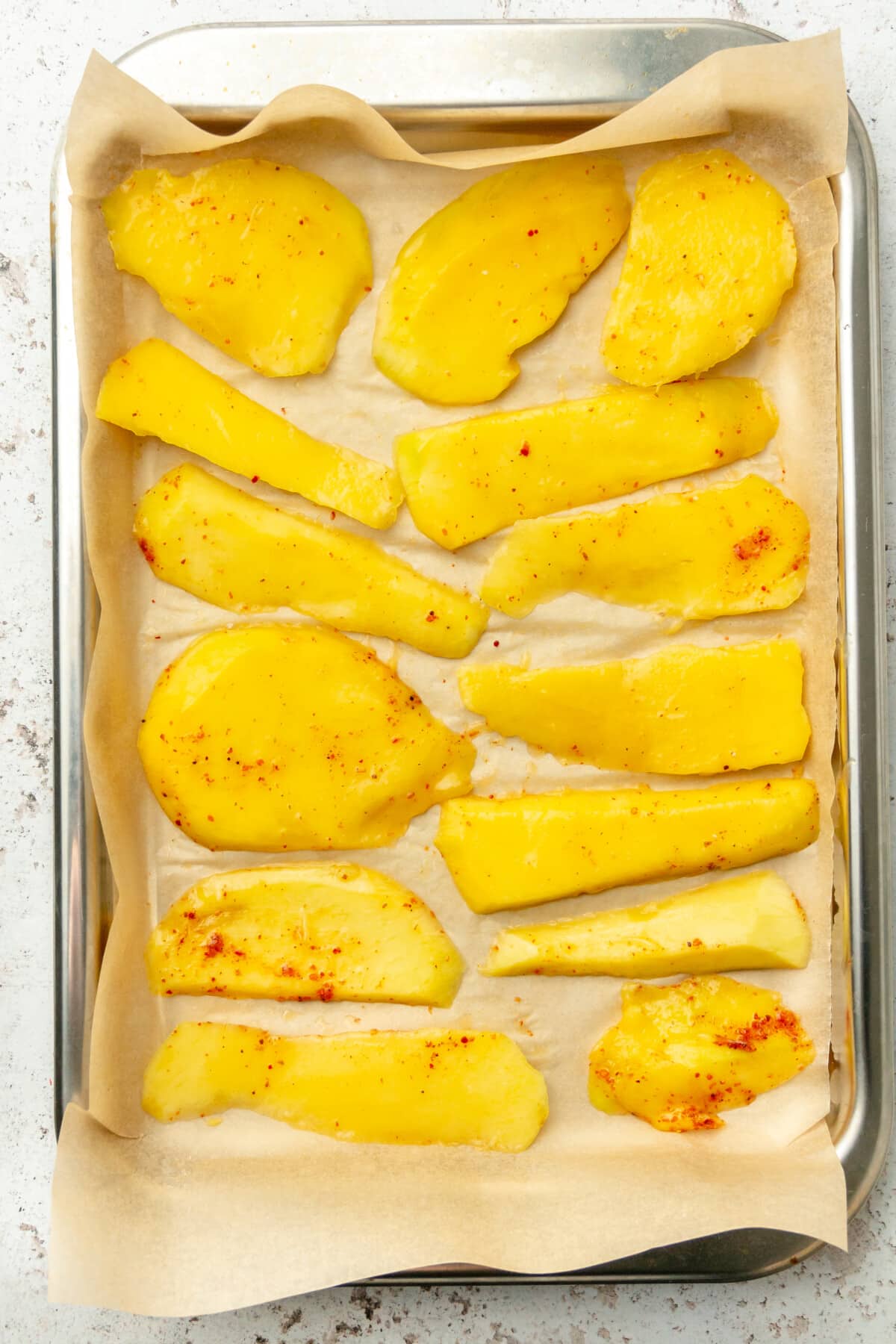 Slices of chili coated mango sit on a lined baking tray on a light grey colored surface.