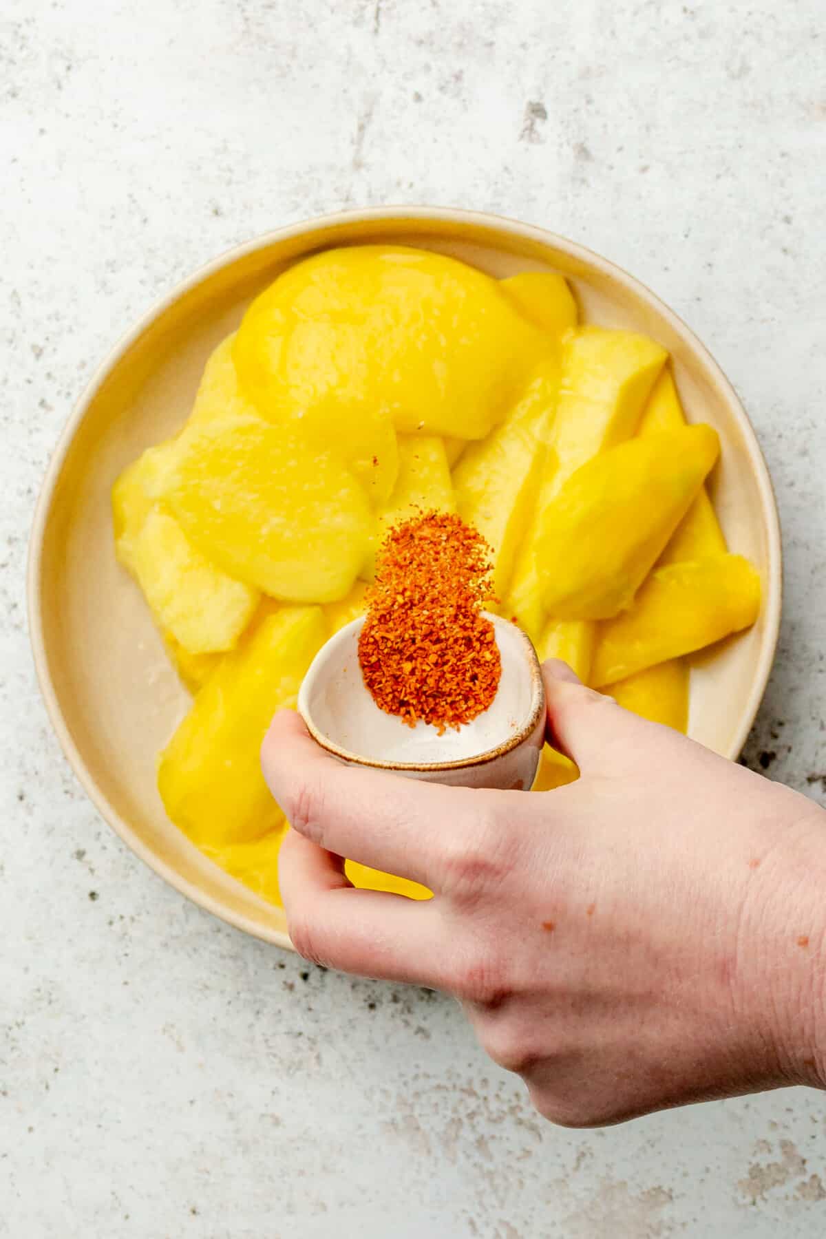 Slices of mango sitting in a shallow bowl have chili tossed over whilst sitting on a light grey colored surface.