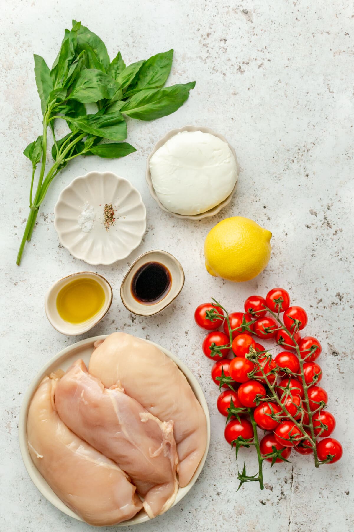 Ingredients for caprese chicken sit in a variety of bowls on a light grey surface.