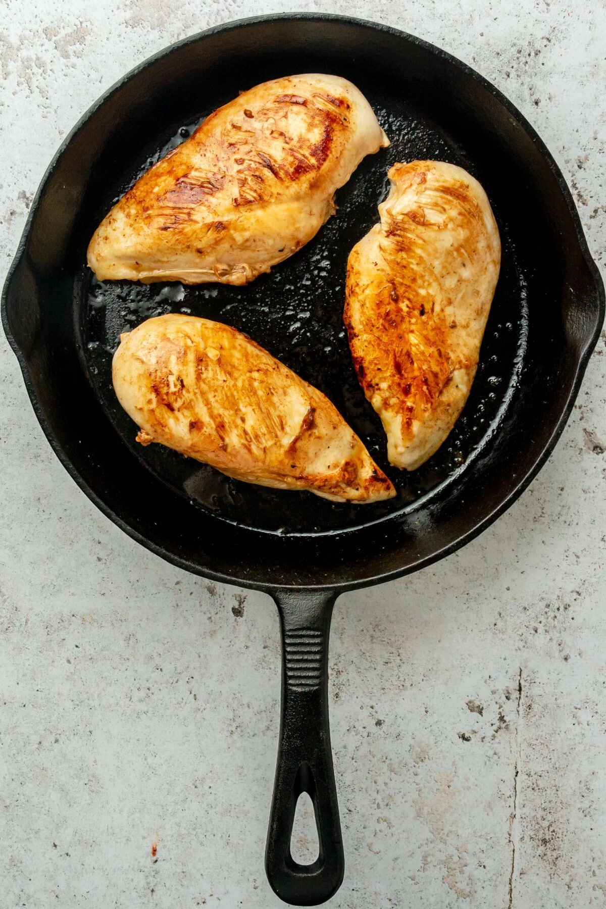 Browned chicken breasts sit in a cast iron skillet on a light grey surface.