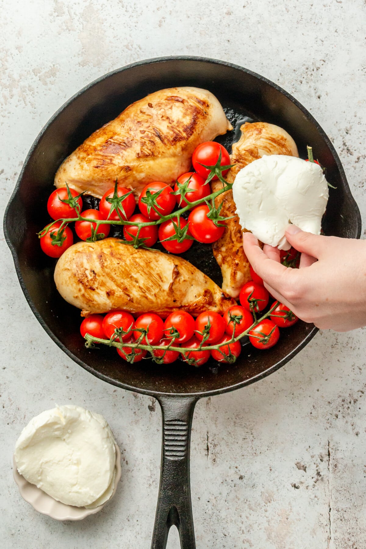 Slices of mozzarella are placed on top of cooked chicken in a cast iron skillet on a light grey surface.