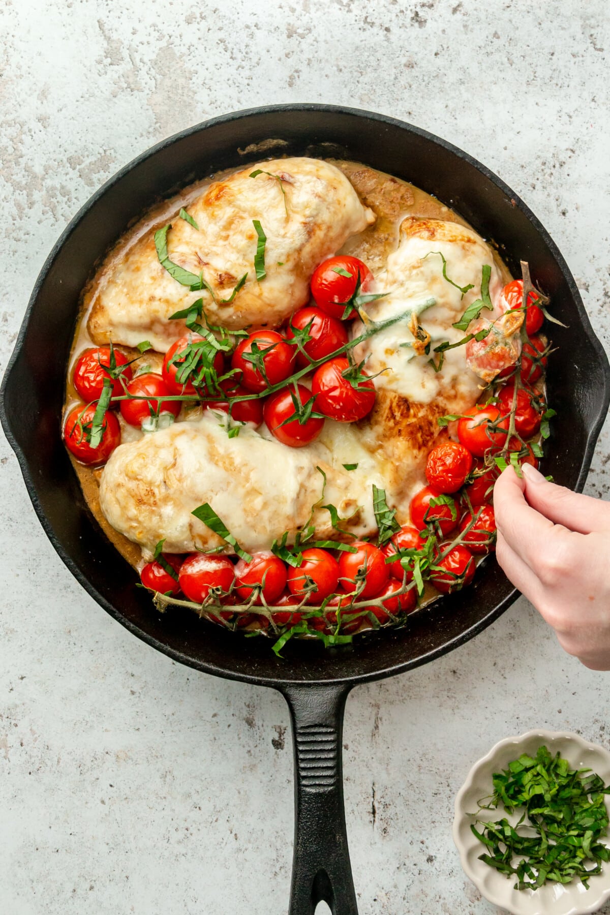 Finely chopped basil is tossed over baked caprese chicken in a cast iron skillet on a light grey surface.
