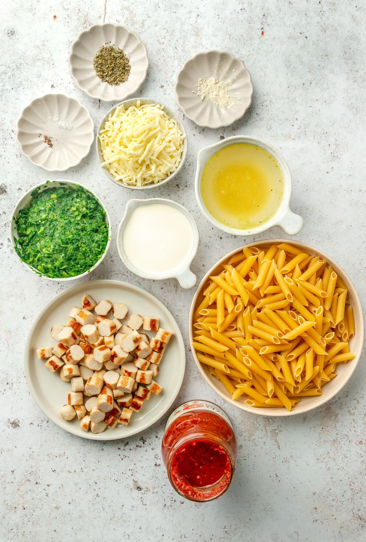 Ingredients for easy no boil pasta bake are laid out in a variety of small plates and bowls on a light grey colored surface.