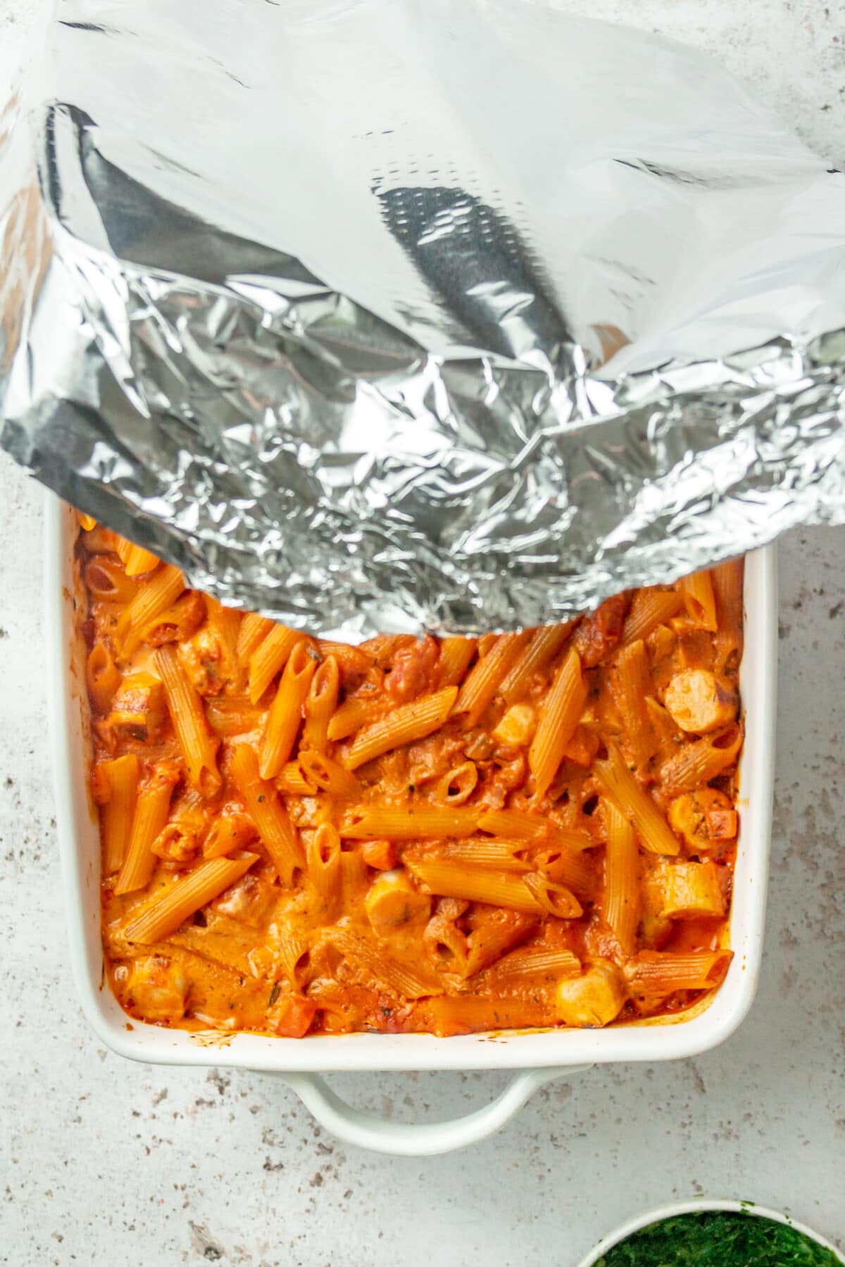 Aluminium foil is pulled off no boil pasta bake in a baking dish on a light grey colored surface.